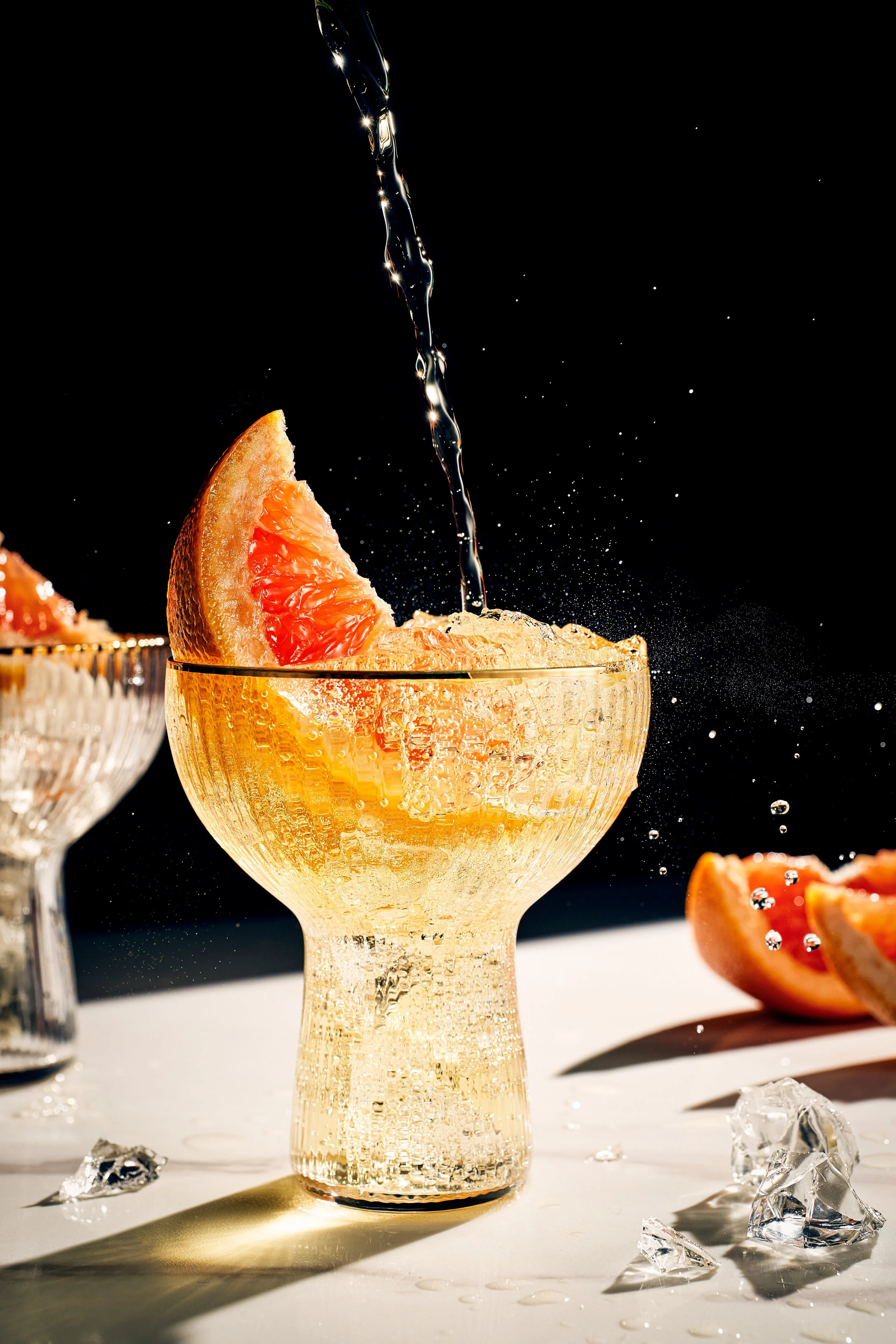 A glass goblet with a textured surface holding a grapefruit wedge, with water being poured into the glass, causing bubbles and splashes, on a white surface with scattered ice cubes and grapefruit slices, against a black background.
