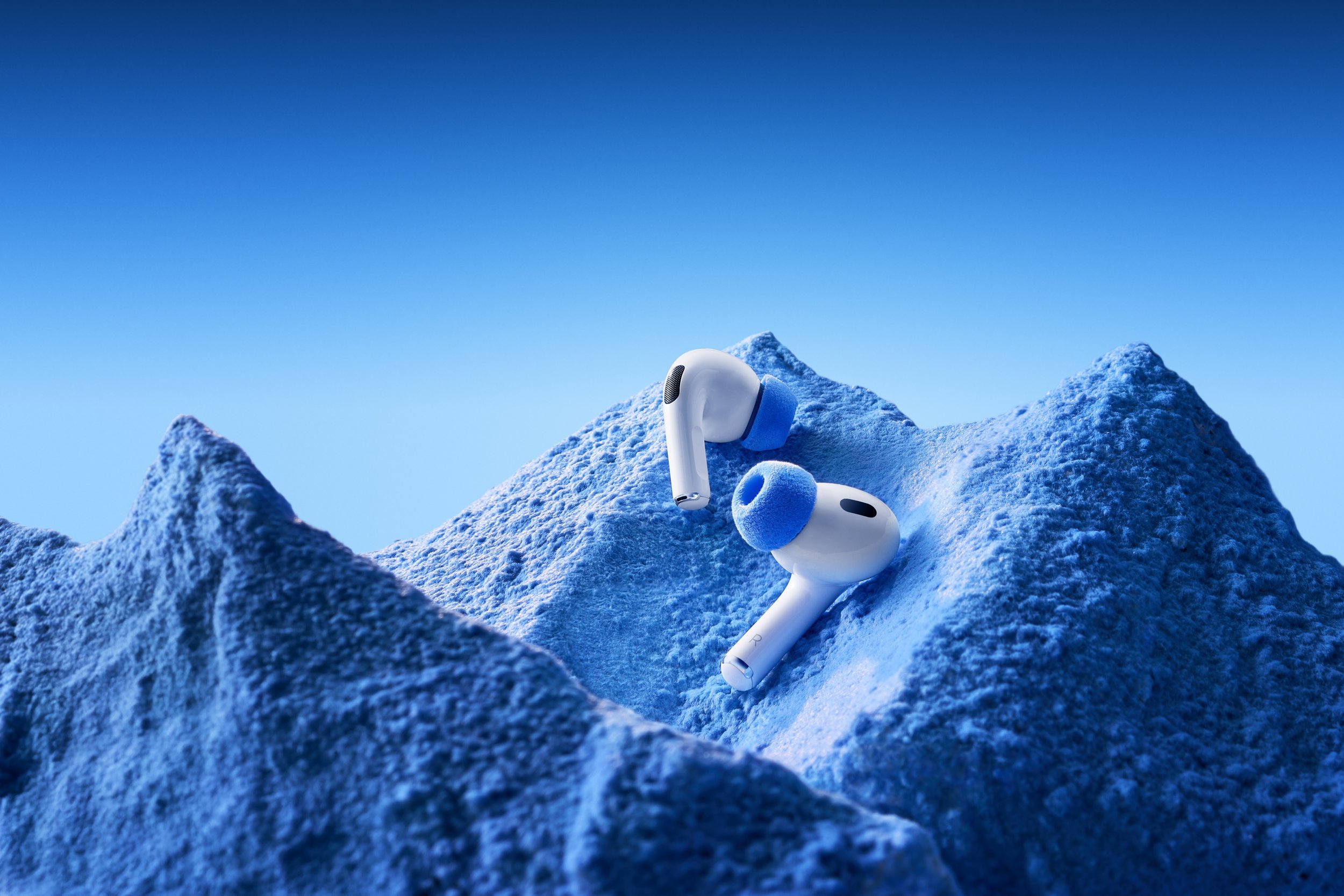 White wireless earbuds with blue ear tips resting on snowy mountain peaks under a clear blue sky.