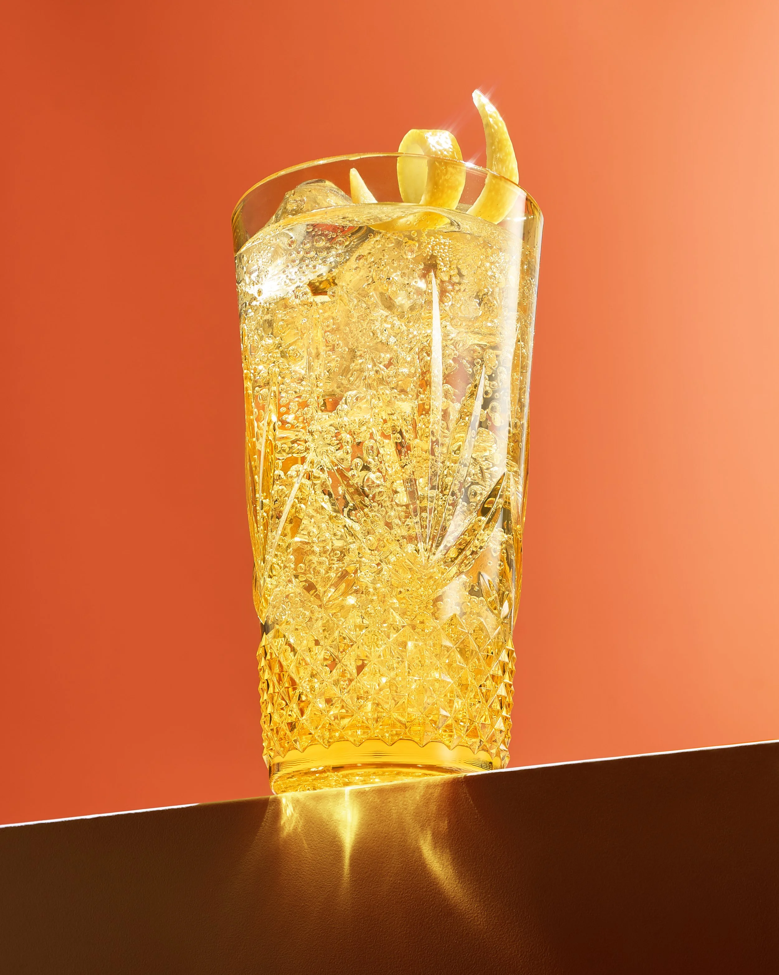 A tall glass filled with a fizzy, yellow-colored beverage, garnished with lemon wedges, with ice cubes visible inside against an orange background.