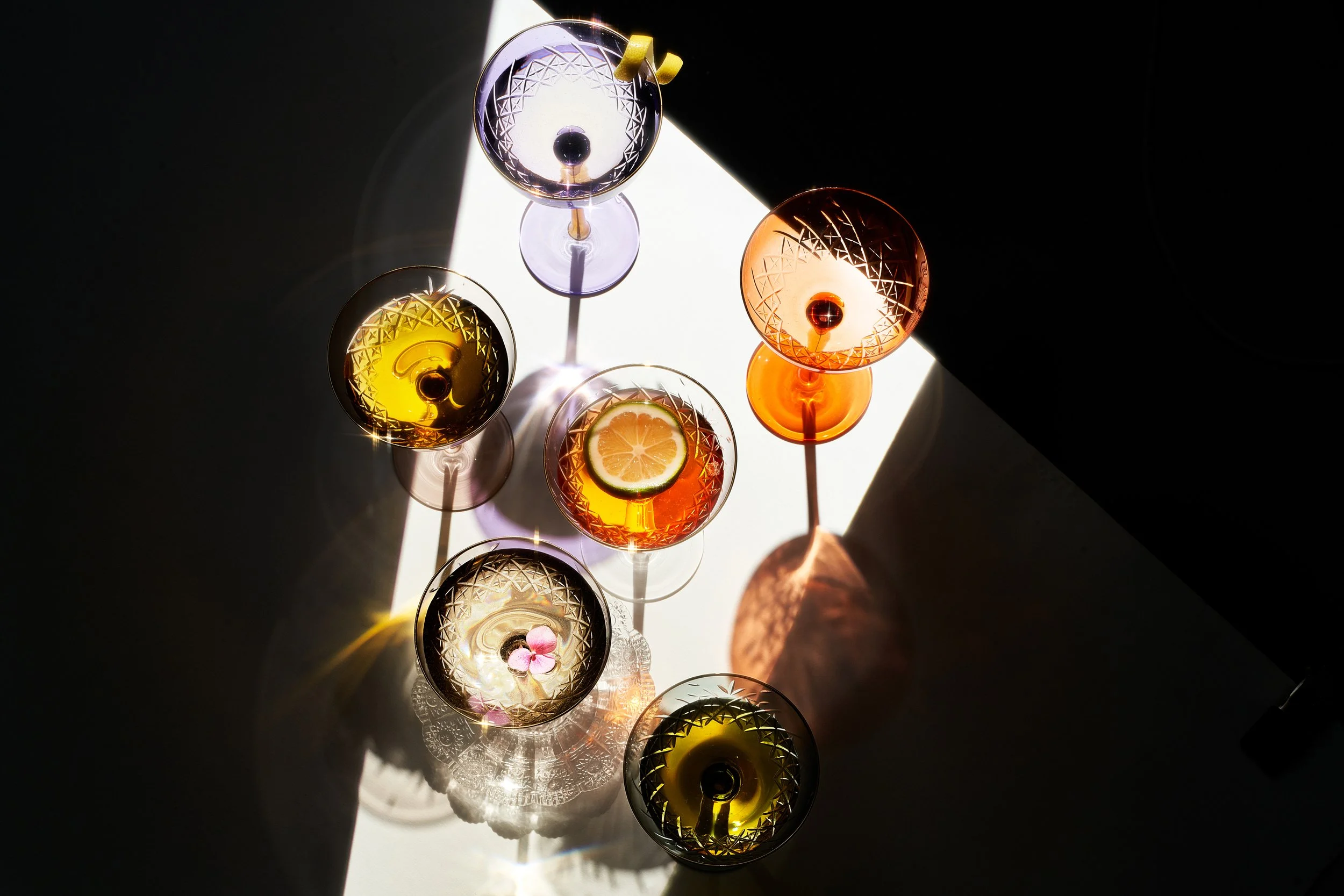 Top-down view of seven colorful cocktail glasses with geometric patterns, some garnished with lemon slices, arranged on a white surface in a triangular formation with shadows cast in different directions. low key, high contrast