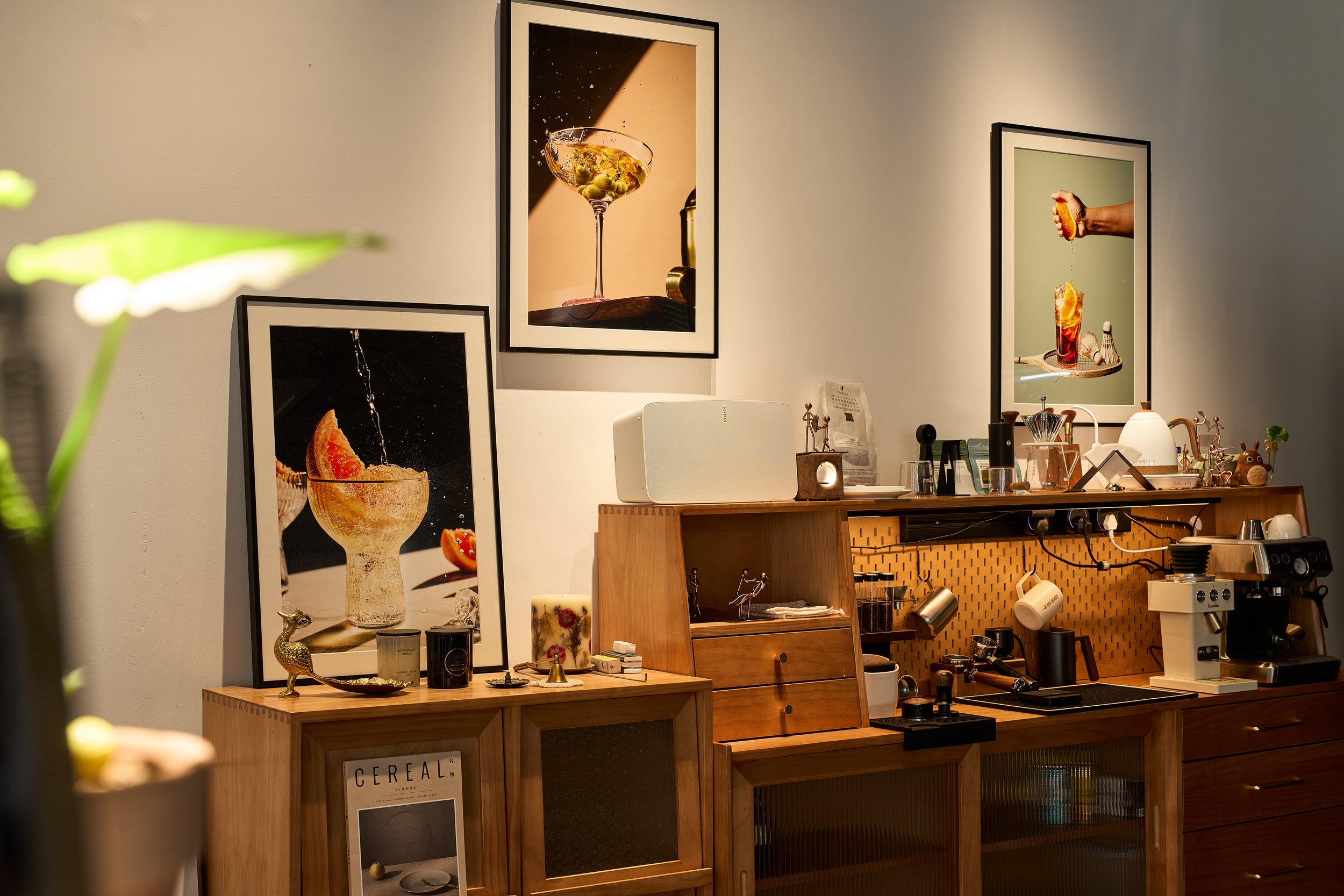 Interior of a cafe or kitchen with framed food and drink photographs on the wall, a wooden counter with kitchen utensils, a coffee machine, and decorative items， coffee corner