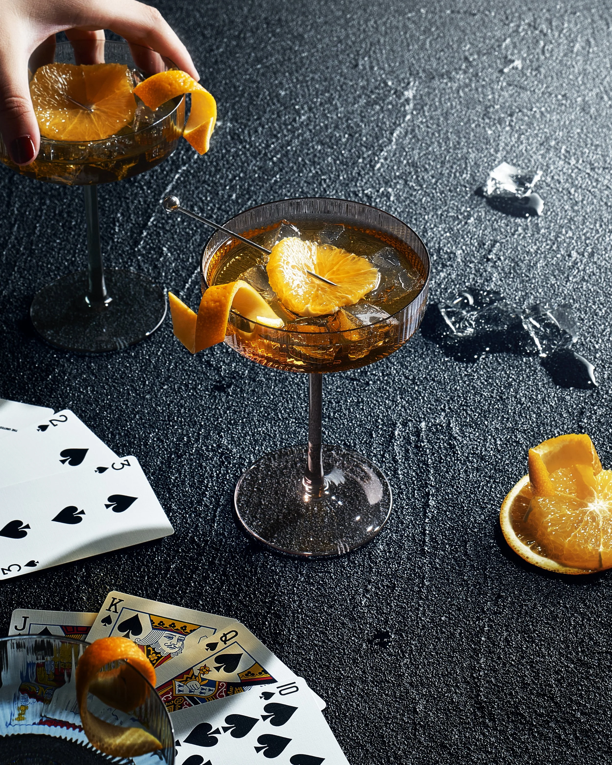Two cocktails garnished with orange slices, with a deck of playing cards and ice cubes on a textured black surface.