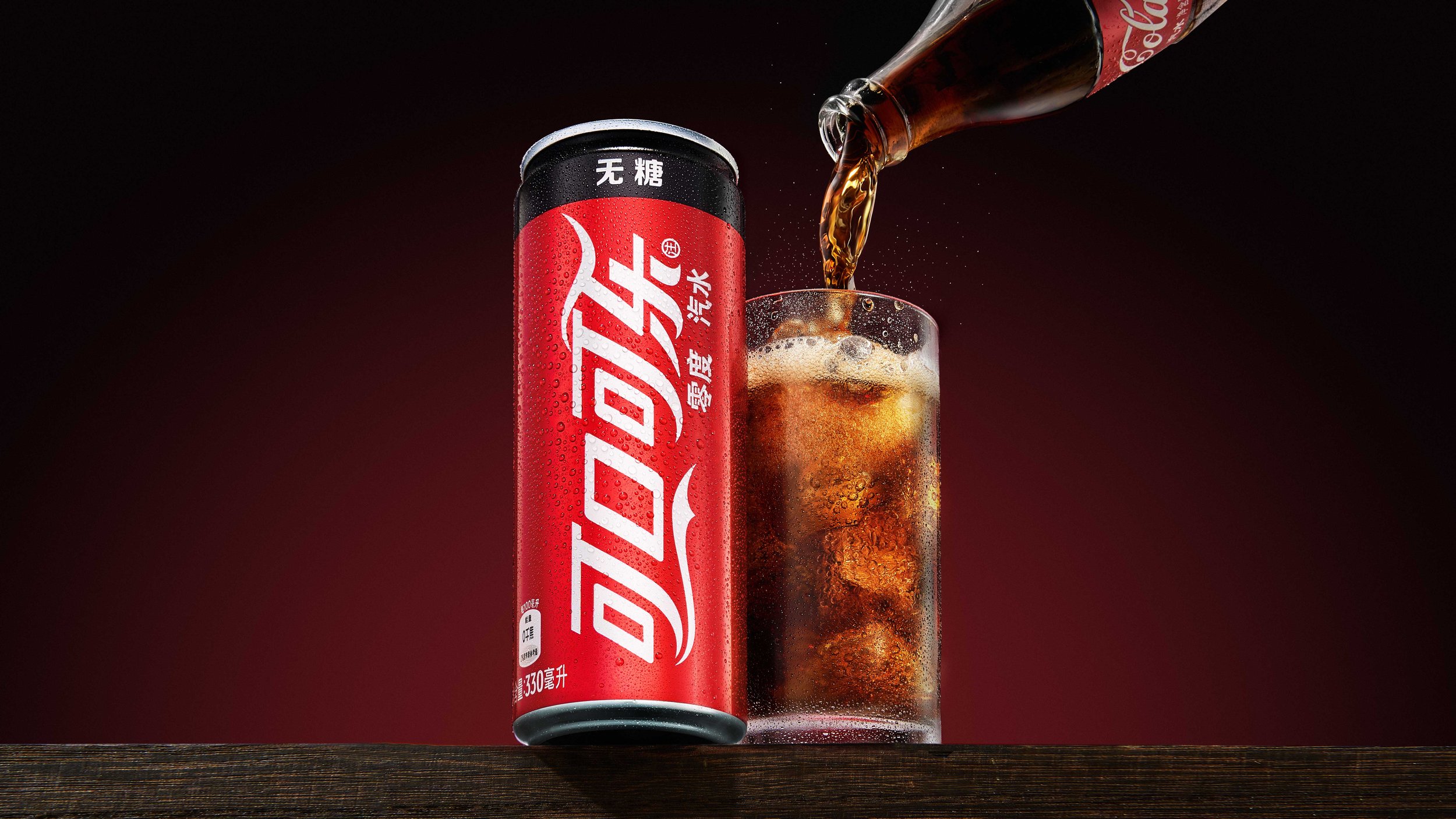 A can of Coca-Cola with Asian characters stands beside a glass filled with ice and Coca-Cola, with more Coke being poured into it from the bottle, against a dark red background.