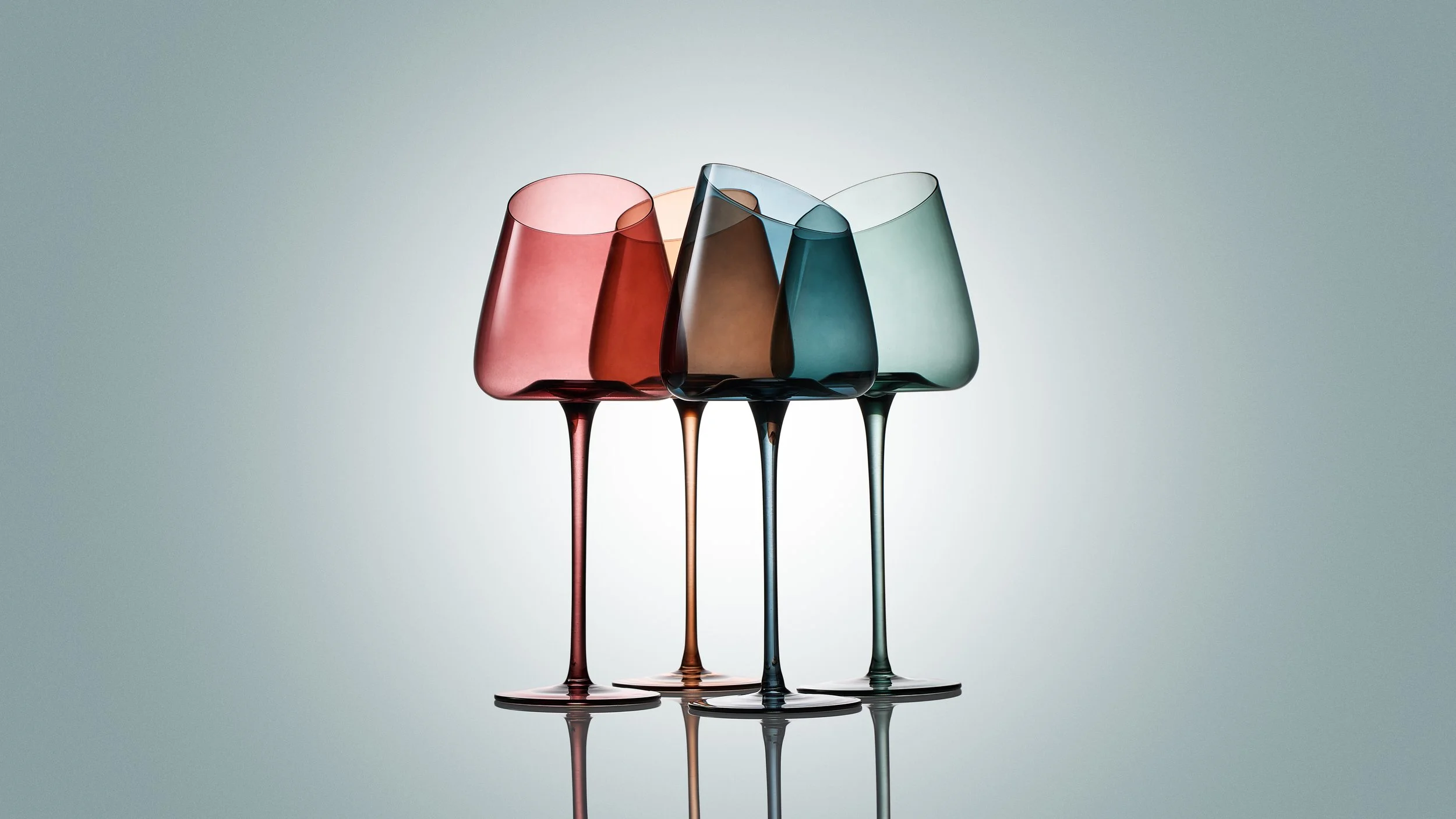 Four colorful wine glasses with long stems, arranged in a row against a neutral background. Each glass is a different color: red, orange, blue, and green.