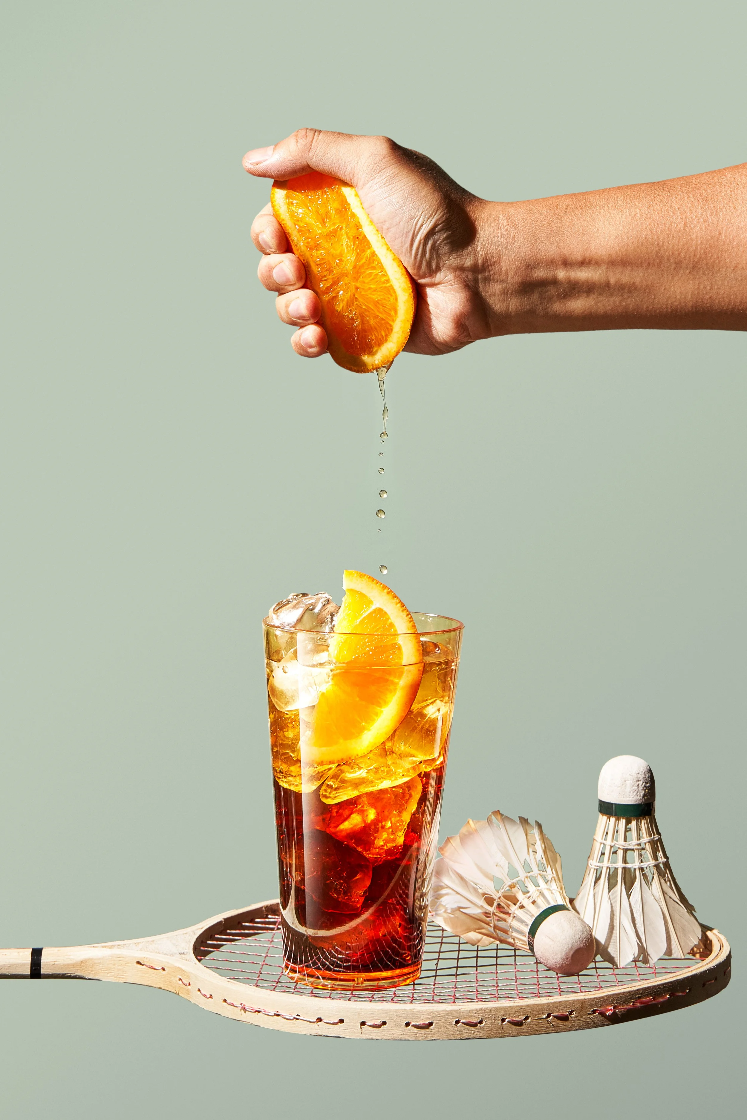 A hand squeezing an orange slice over a glass of iced tea with orange slices and ice cubes, with badminton shuttlecocks on a racket below.