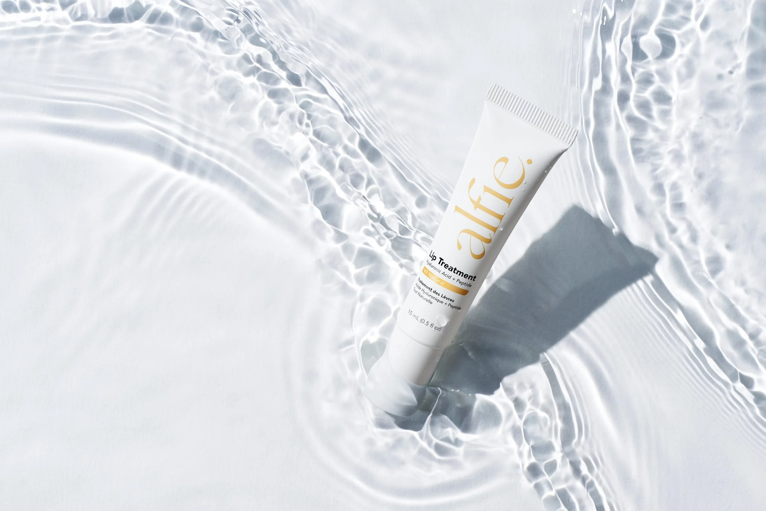 A white tube of ALFA Lip Treatment with hyaluronic acid and peptide, partially submerged in water, creating ripples with sunlight reflections.