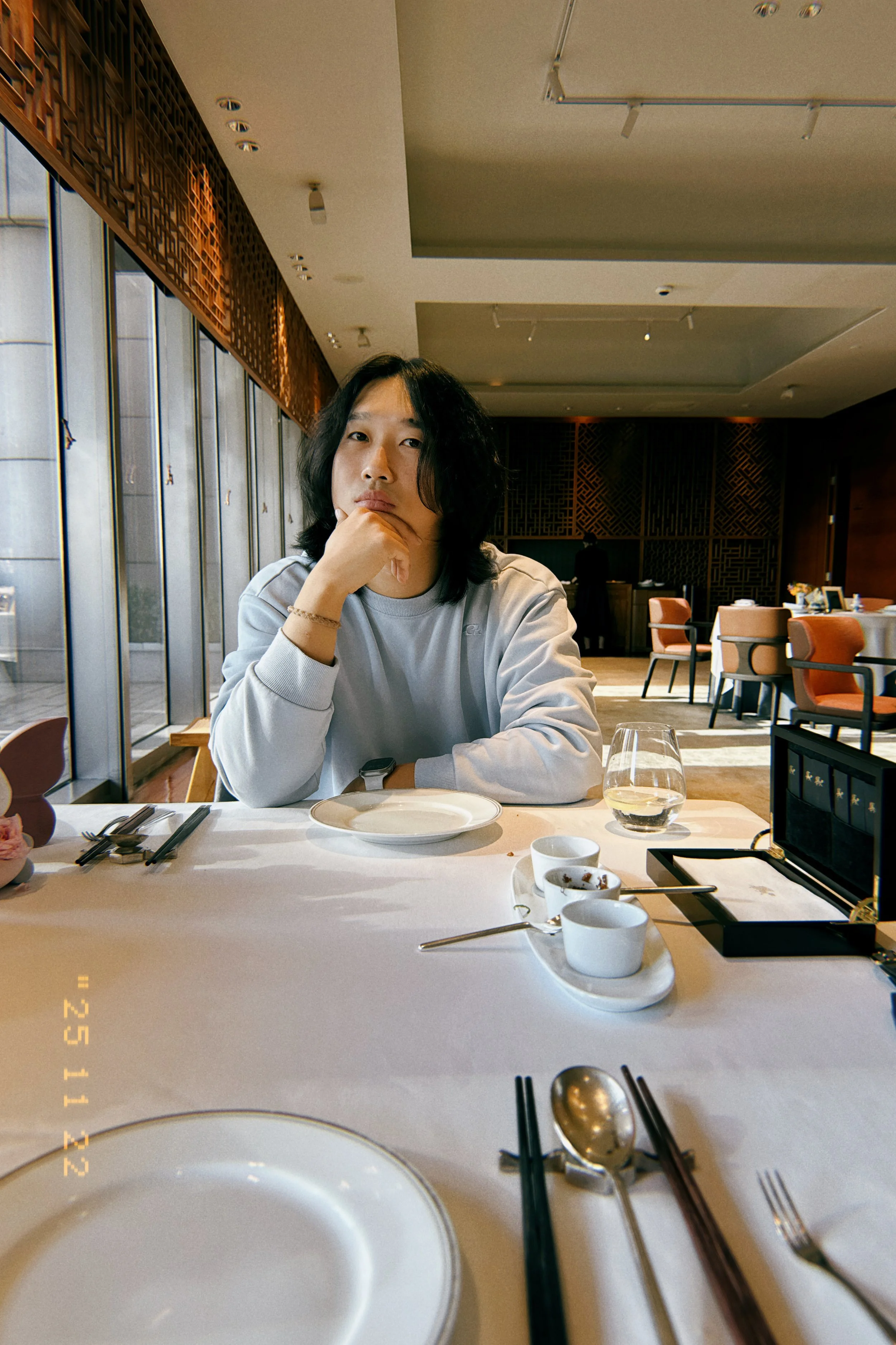 A person with shoulder-length black hair sitting at a restaurant table, resting their chin on their hand, with empty plates, glasses, and utensils on the table, and large windows allowing natural light.