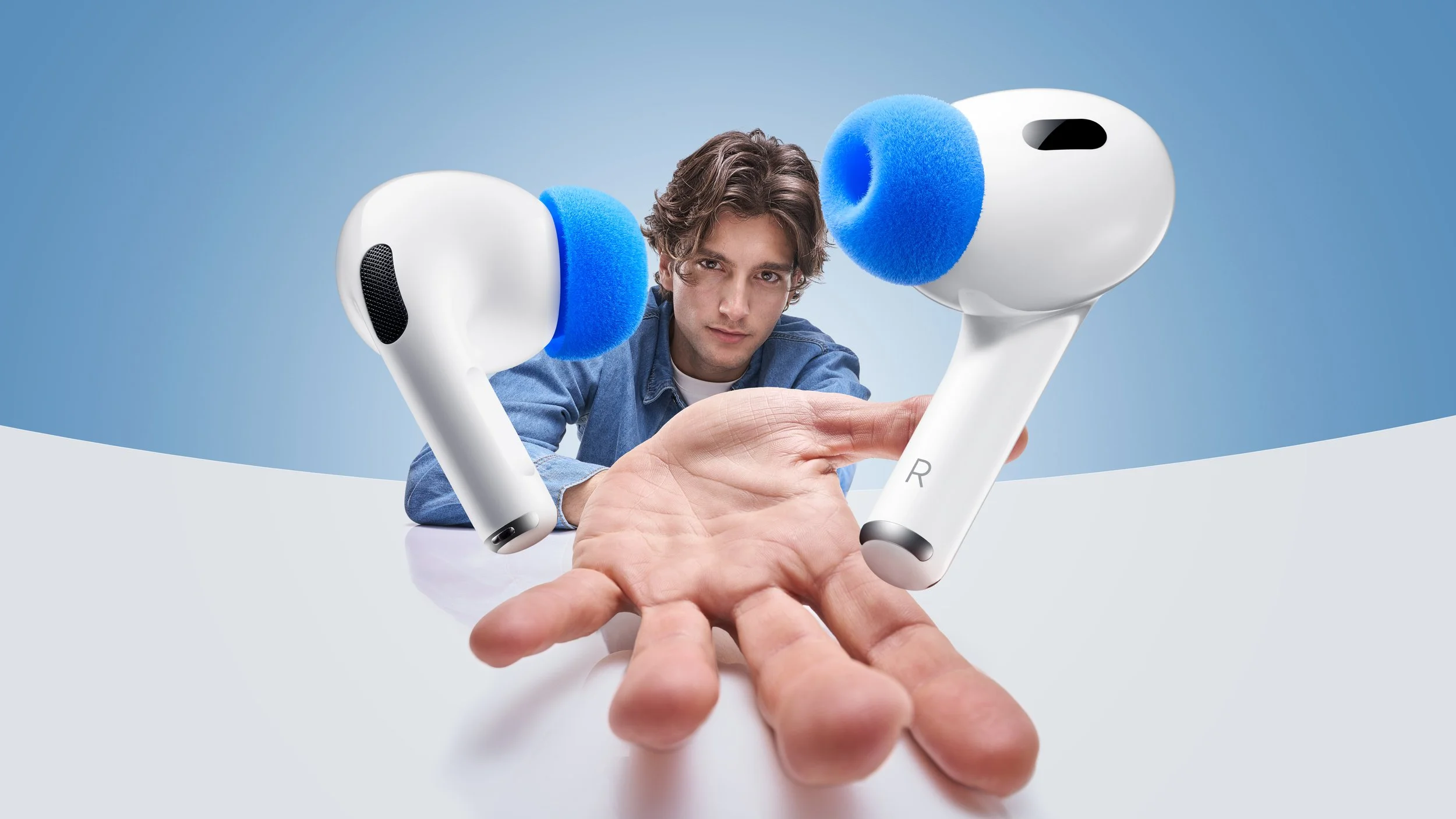 A young man with brown hair in a denim jacket and white shirt is reaching out with a hand and holding a white wireless earbuds case with blue ear tips, with the earbuds floating near his face against a light blue background.