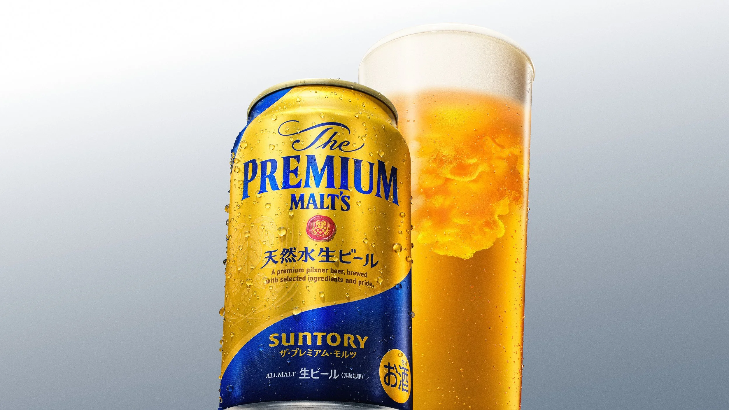 A can of Suntory The Premium Malt's beer next to a glass filled with beer, showing condensation on the can and beer foam on top of the glass.