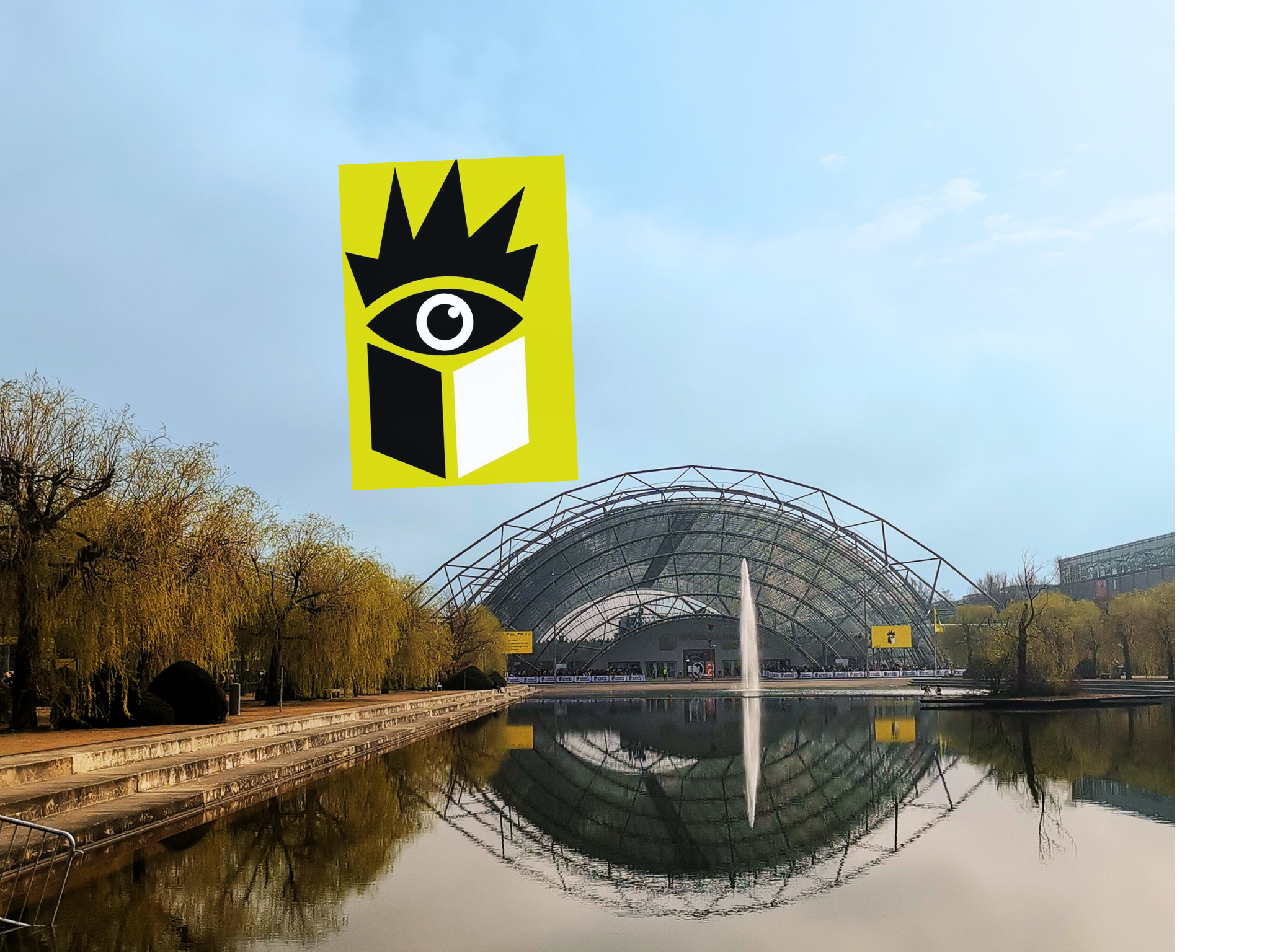 Ein moderner Park mit einem Wasserbecken, einer Springbrunnen und einem gläsernen Bauwerk im Hintergrund, umgeben von Bäumen im Herbst, bei bewölktem Himmel.