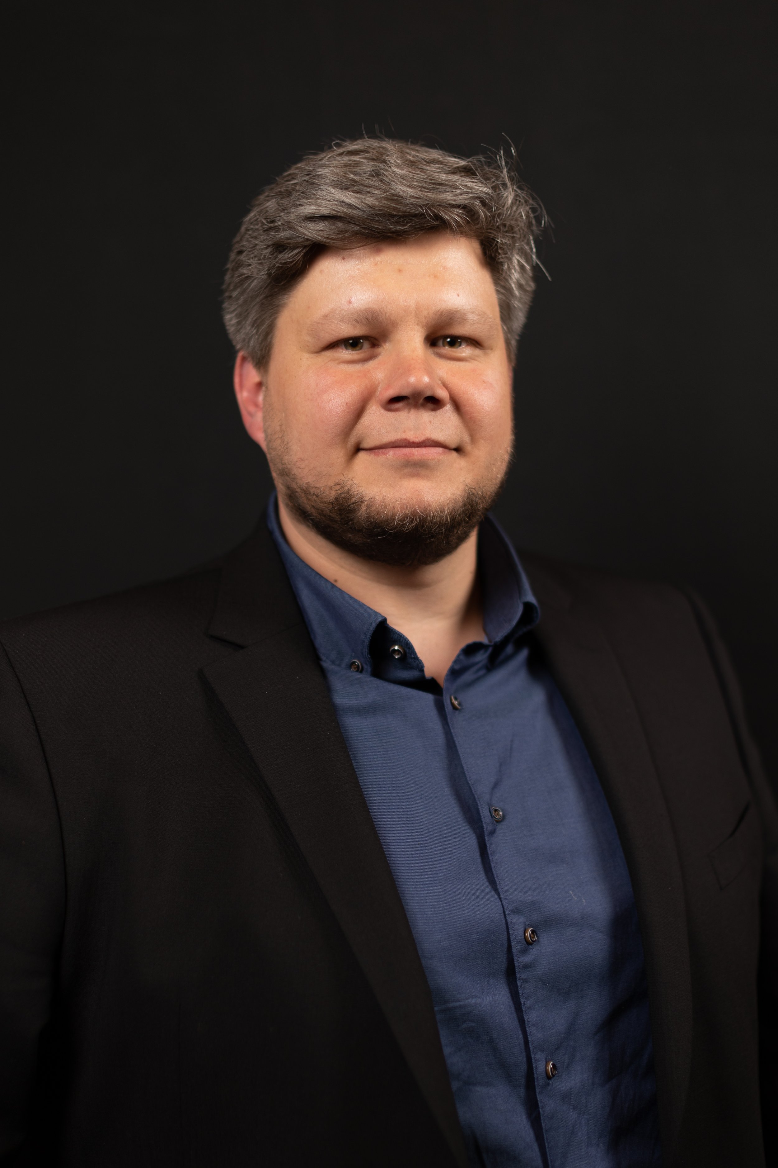 Ein Mann mit grauen Haaren und Bart trägt einen schwarzen Anzug und ein dunkles Hemd, steht vor einem schwarzen Hintergrund.