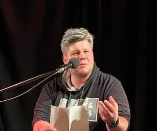 Mann bei einer Lesung, sitzt vor einem Mikrofon, liest aus einem Buch vor einem schwarzen Hintergrund.