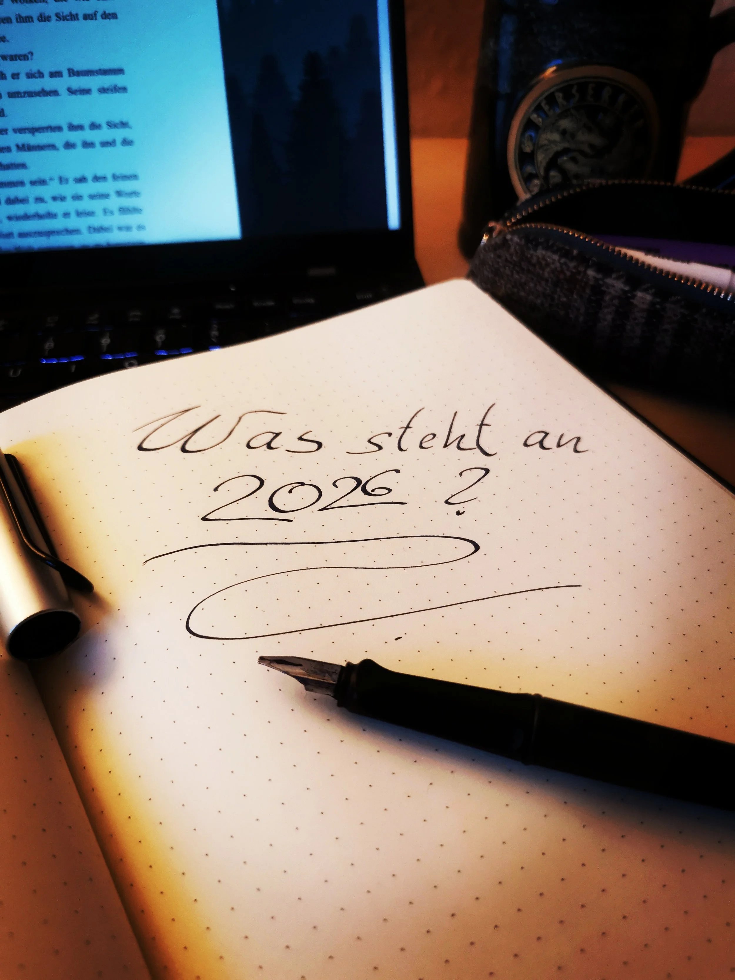 Ein offenes Notizbuch mit der Hand geschriebenen Frage ‚Was steht an 2026?‘, ein schwarzer Kugelschreiber, eine Tasche, eine Tasse und ein Laptop. Die Umgebung ist dunkel.