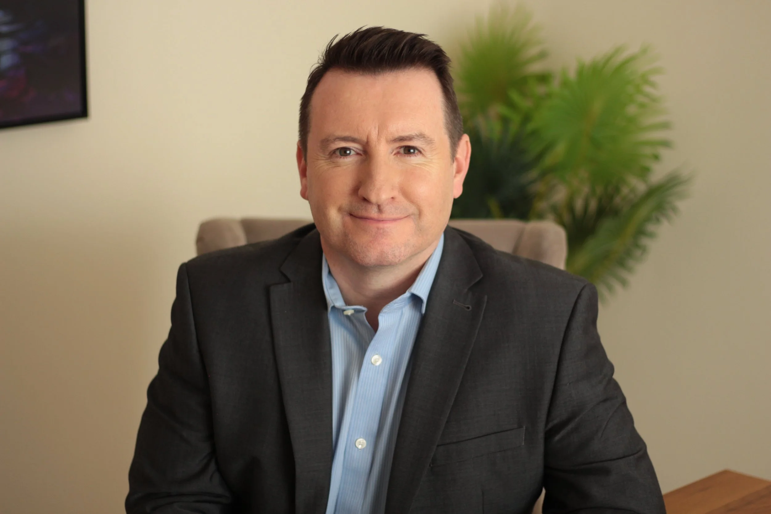 A man with short dark hair and a light complexion, wearing a dark suit jacket and a light blue shirt, sitting in an office with a beige wall and a green leafy plant in the background.