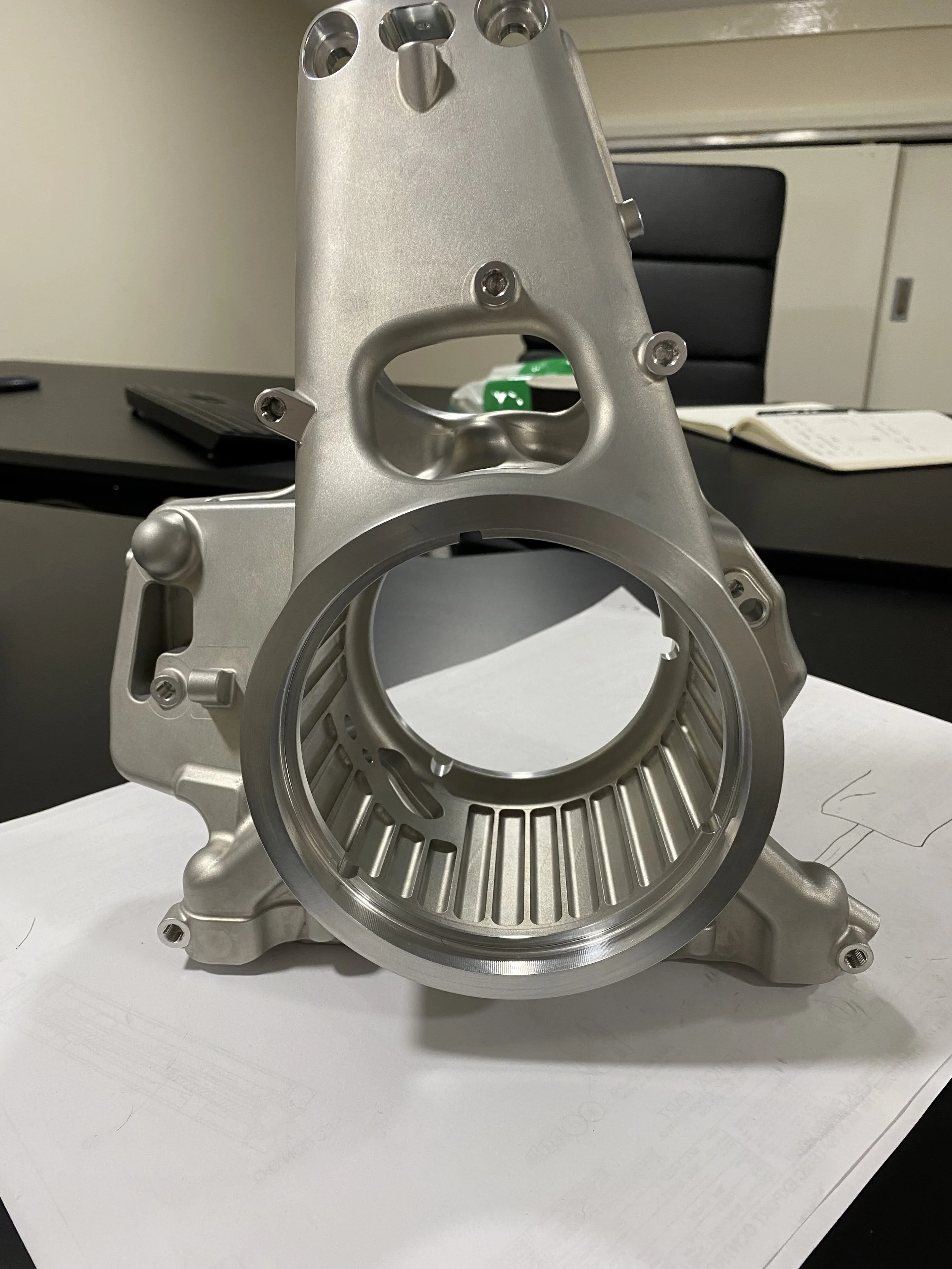 A metallic mechanical component, possibly part of a machine or engine, is placed on a white surface with a black chair, a notebook, and some green items in the background.