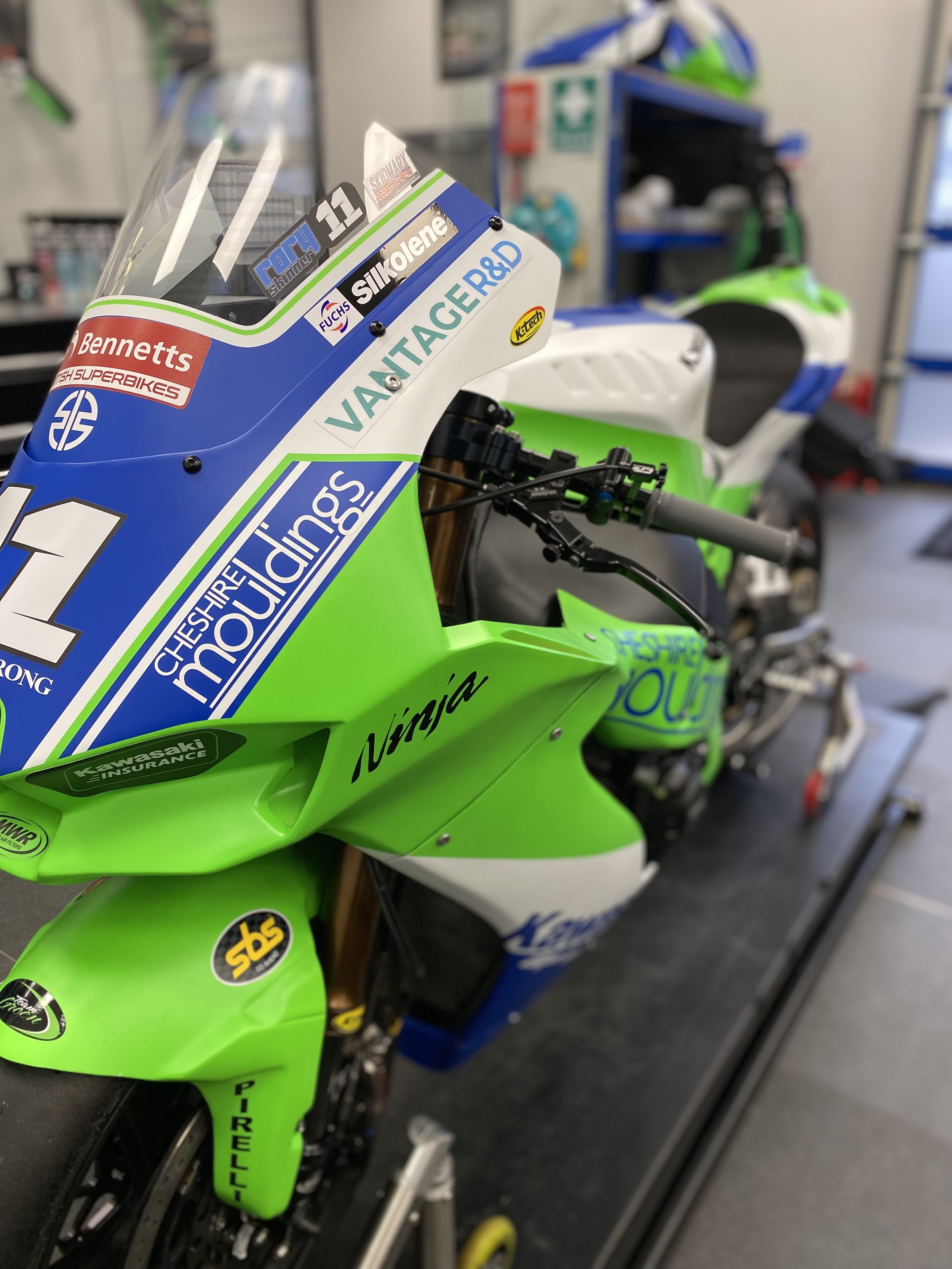 Close-up of a bright green and blue Kawasaki racing motorcycle with various sponsor stickers, placed on a maintenance stand in a workshop.