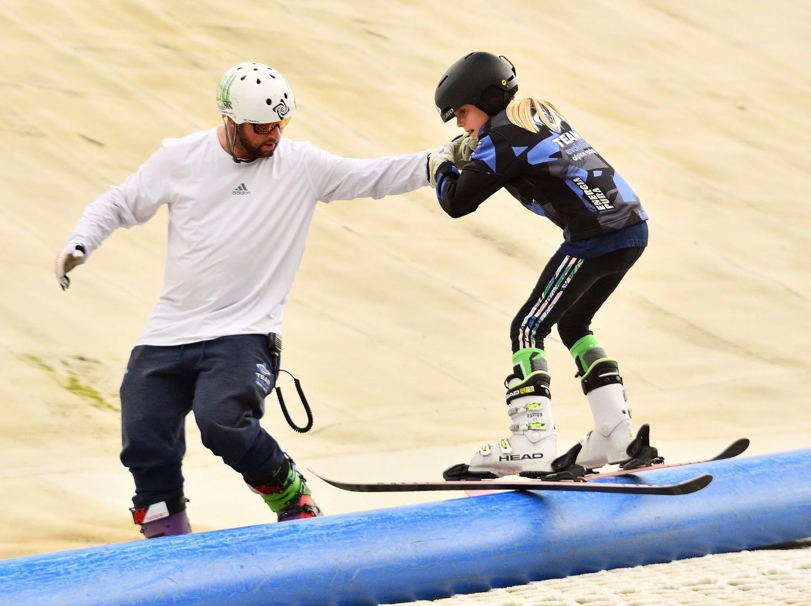 A coach helps a young female skier ride a rail at a dry ski slope