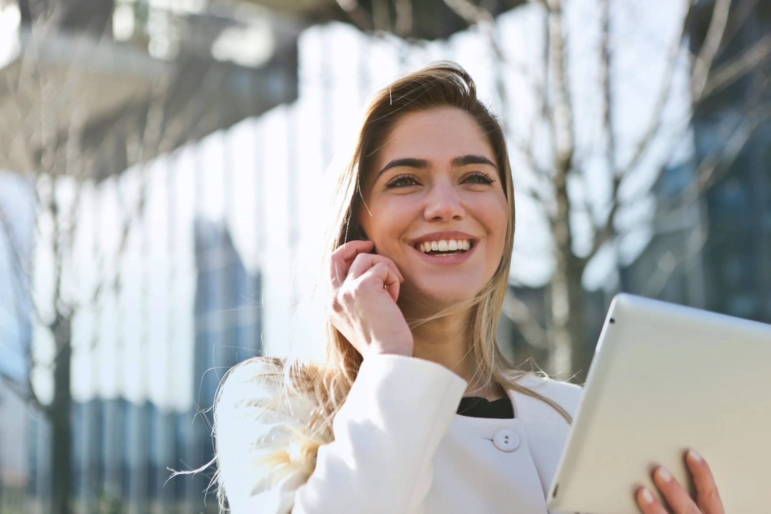 A young woman smiling outdoors, holding a tablet and talking on her phone, with bare trees and modern buildings in the background.