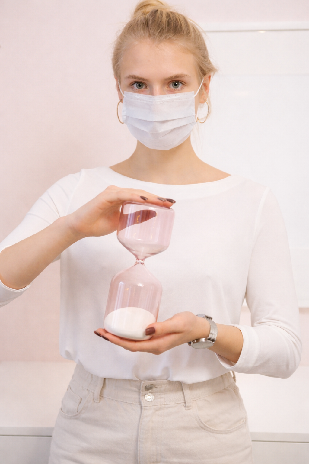 A young woman with blonde hair, wearing a white face mask and white top, holds an hourglass with pink glass and white sand, with the sand flowing from the upper to the lower chamber.