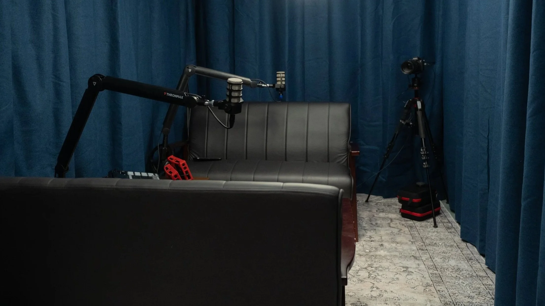 Podcast studio with blue curtains and two microphones, two black sofas and a camera