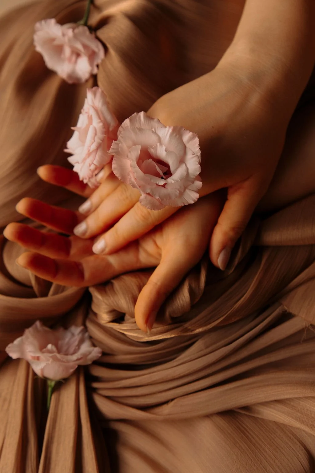 Une main posée sur une autre avec des fleurs roses enroulées autour des doigts et d'autres fleurs posées sur un tissu ou un vêtement en dessous.
