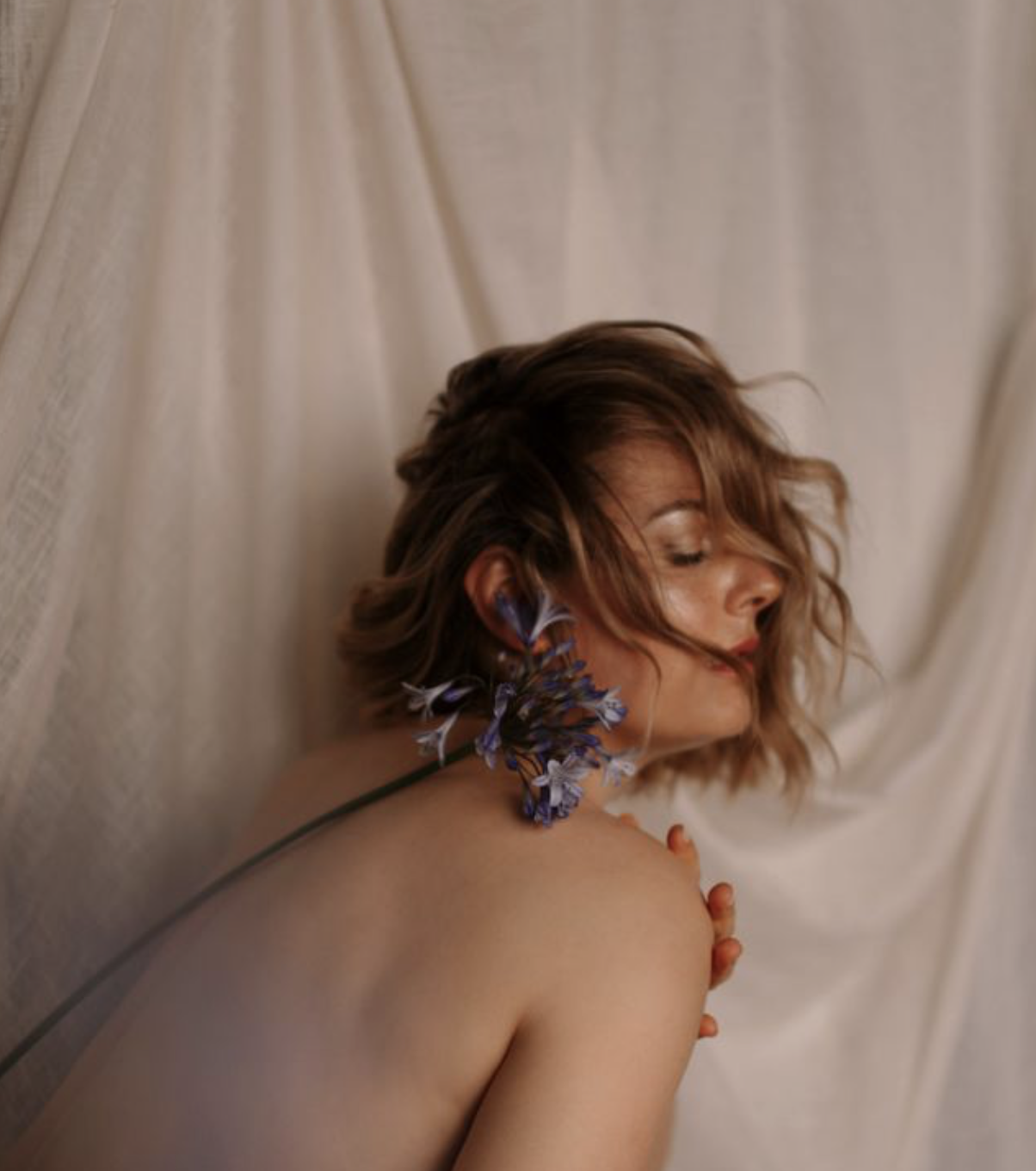 Femme nue posant contre un mur en tissu beige, portant une grosse boucle d'oreille avec des fleurs violet et blanc.