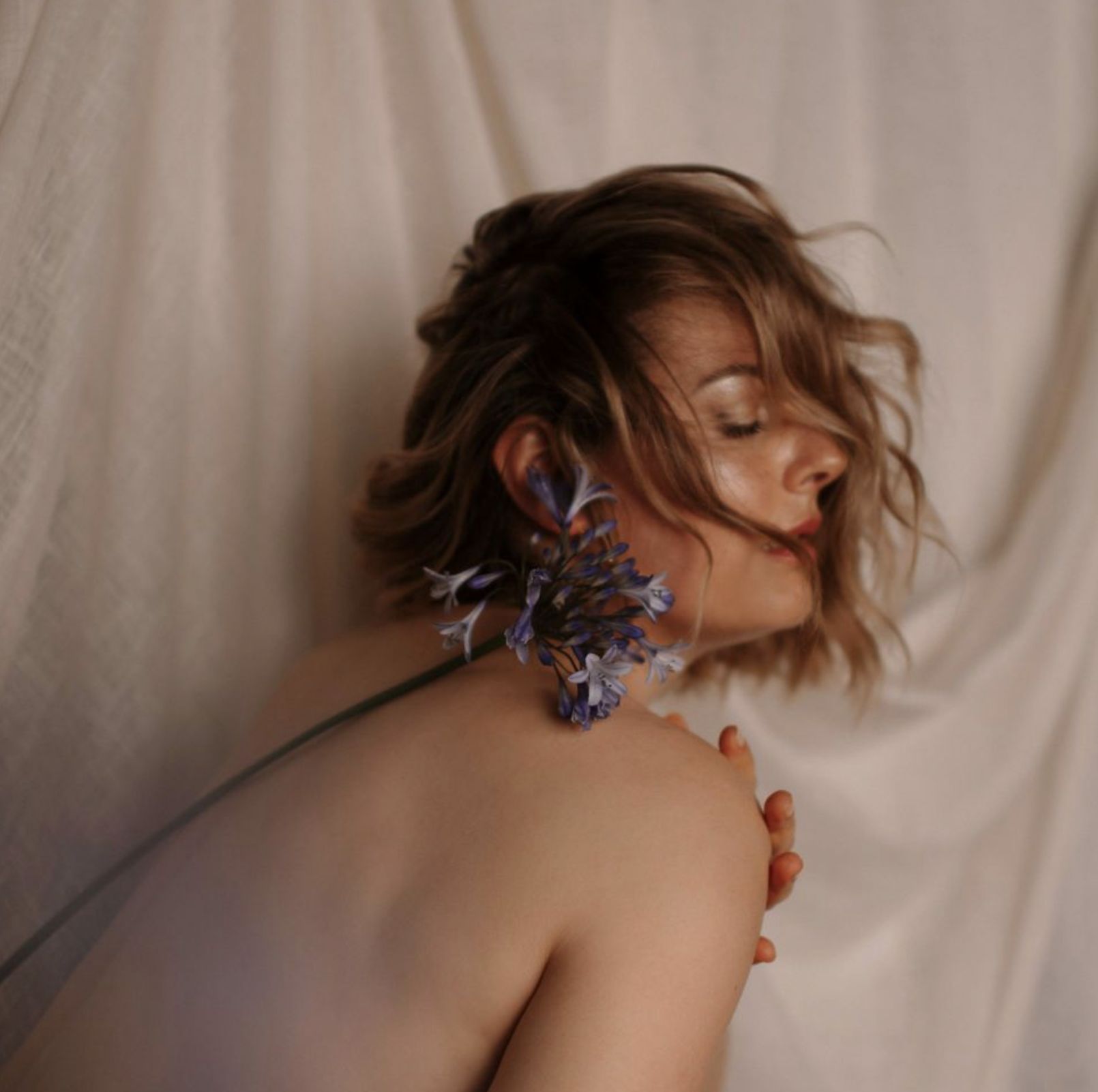 Une femme aux yeux fermés, les cheveux châtains ondulés, repose contre un mur en tissu beige, avec une fleur violette à côté de son visage.