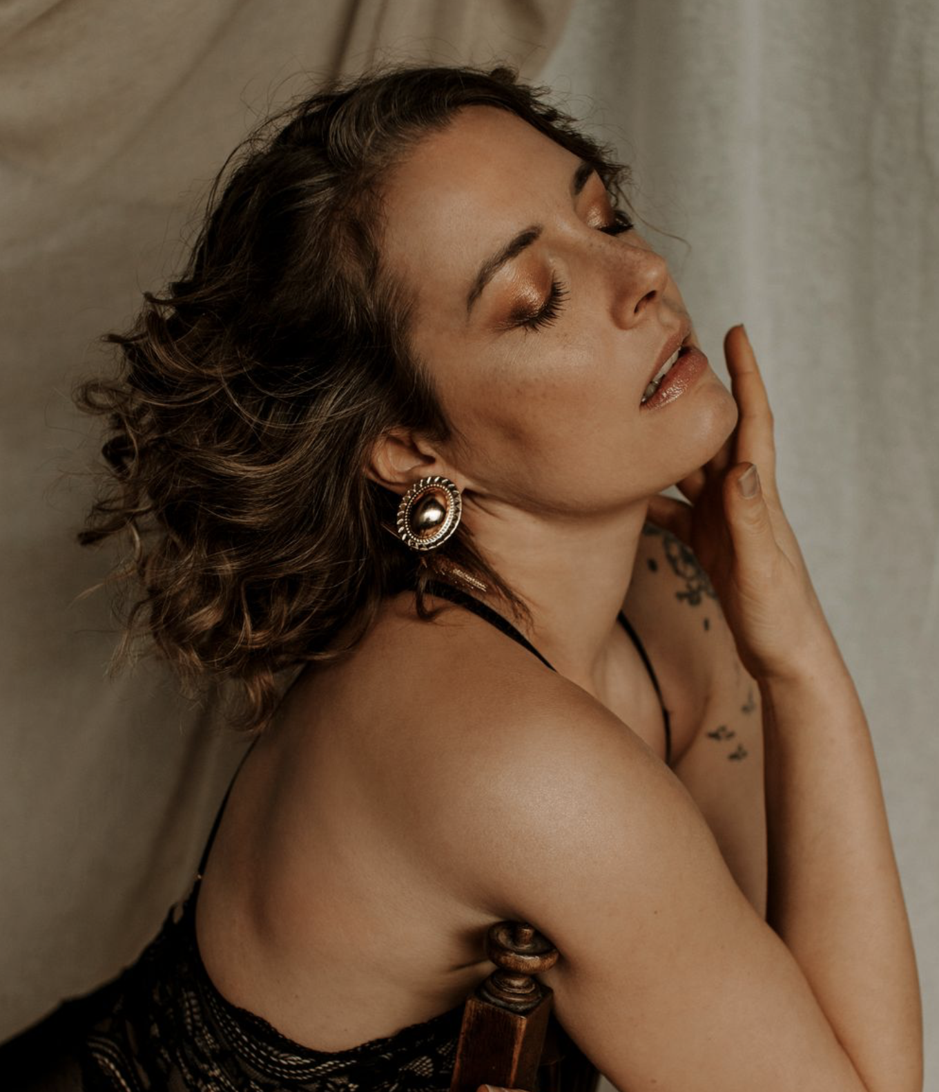 Femme avec des cheveux bouclés, portant un tatouage sur l'épaule, portant une boucle d'oreille et un vêtement noir en dentelle, posant avec les yeux fermés et touchant son visage, dans un décor intérieur