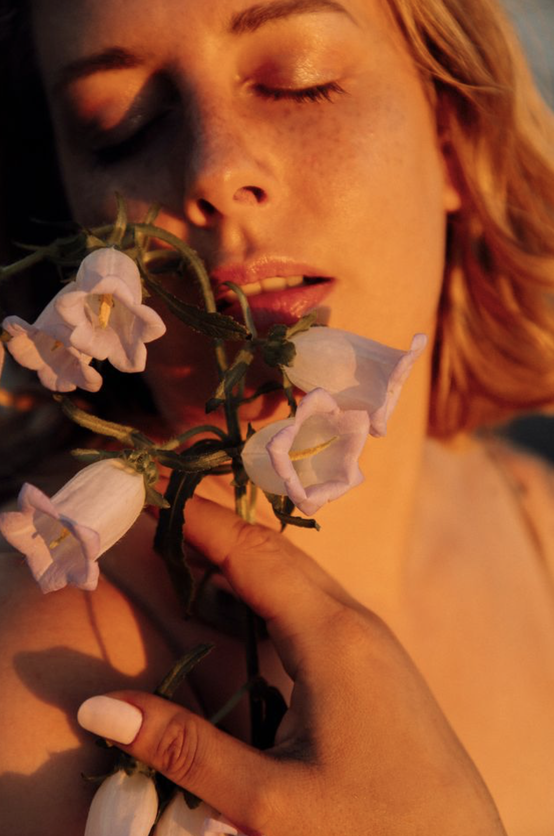 Portrait d'Alice Alma avec les yeux fermés, des lèvres légèrement entrouvertes, tenant un bouquet de fleurs blanches près de son visage, avec un éclairage chaud et doux.