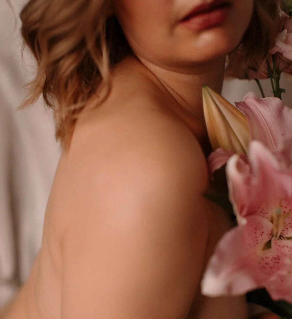 Un gros plan de l'épaule et du cou d'une femme, avec des fleurs roses à côté, lèvres partiellement visibles, cheveux bouclés
