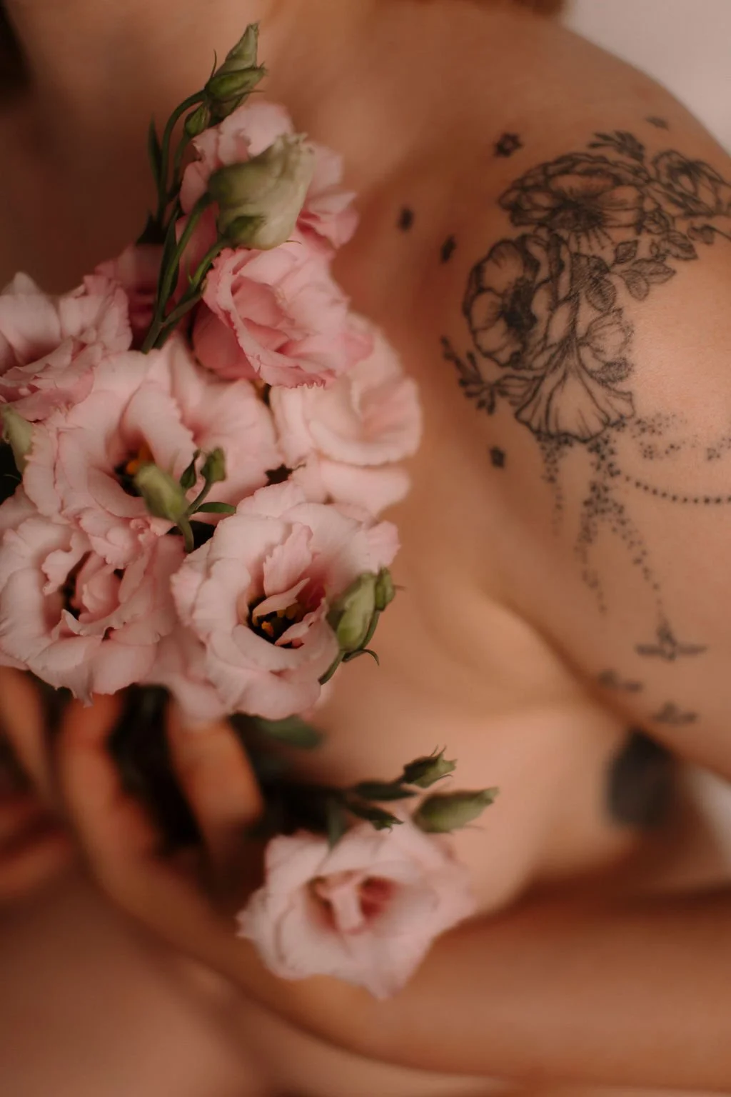 Photo artistique d'une personne nue tenant un bouquet de fleurs roses, avec un tatouage floral noir sur l'épaule.