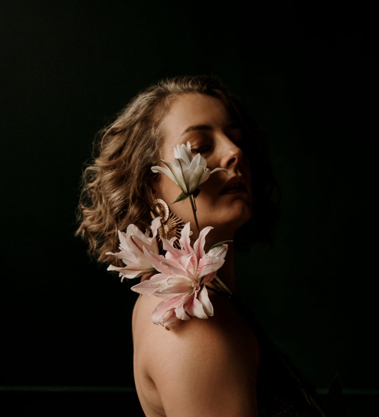 Femme aux cheveux bouclés portant des boucles d'oreilles, avec des fleurs blanches et roses près de son visage et sur son épaule, fond noir.