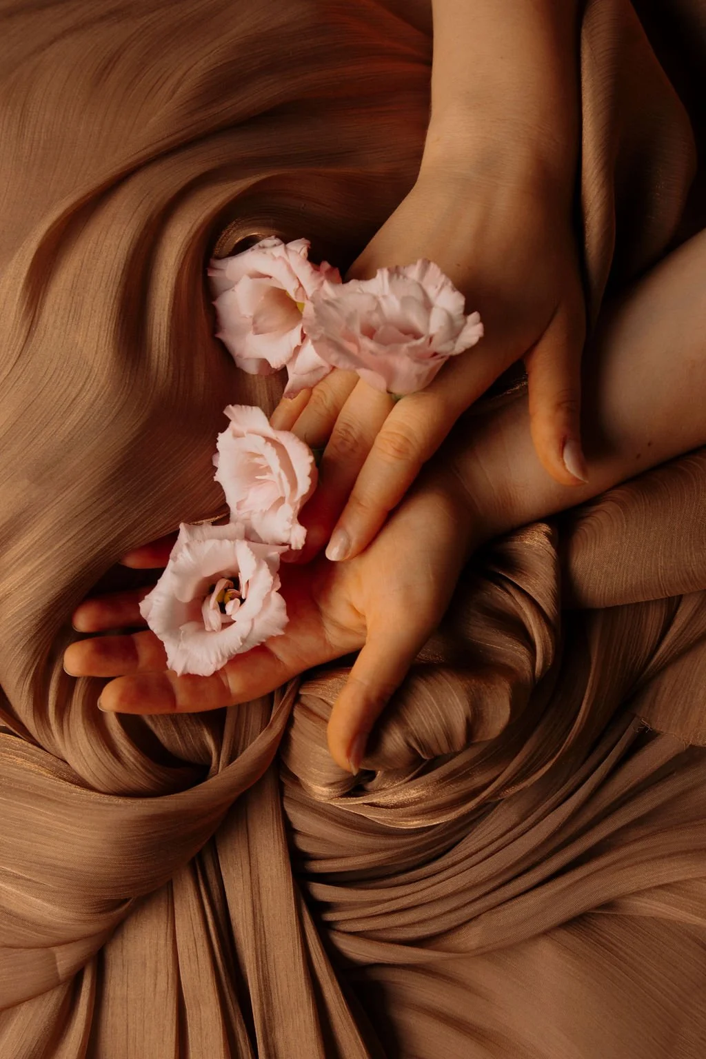 Deux mains délicates, une avec trois fleurs roses, reposent sur un tissu en satin beige avec des plis et des courbes.
