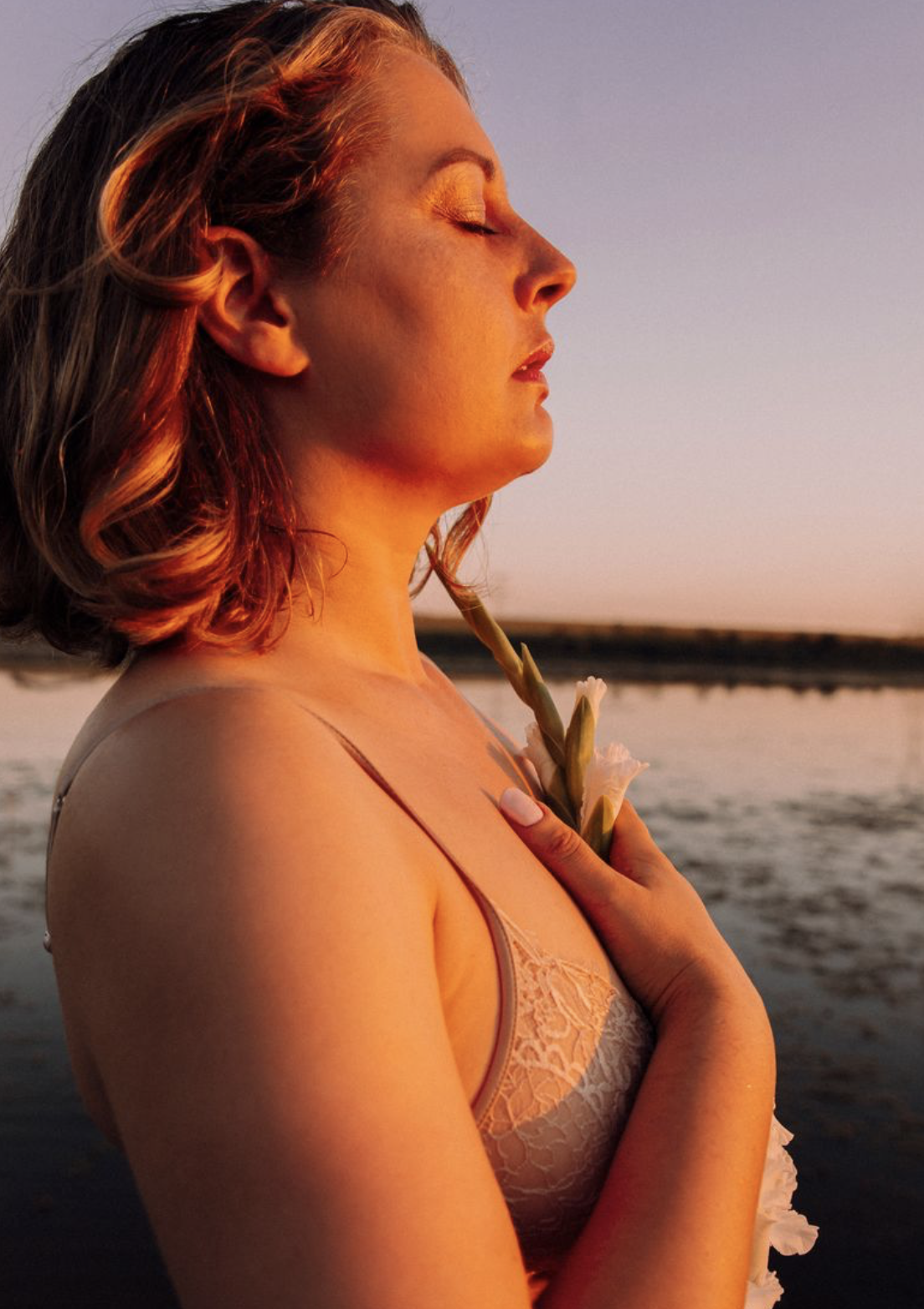Une femme avec des cheveux roux courts, les yeux fermés, tenant une fleur blanche près de la poitrine, silhouette contre un coucher de soleil au bord de l'eau.