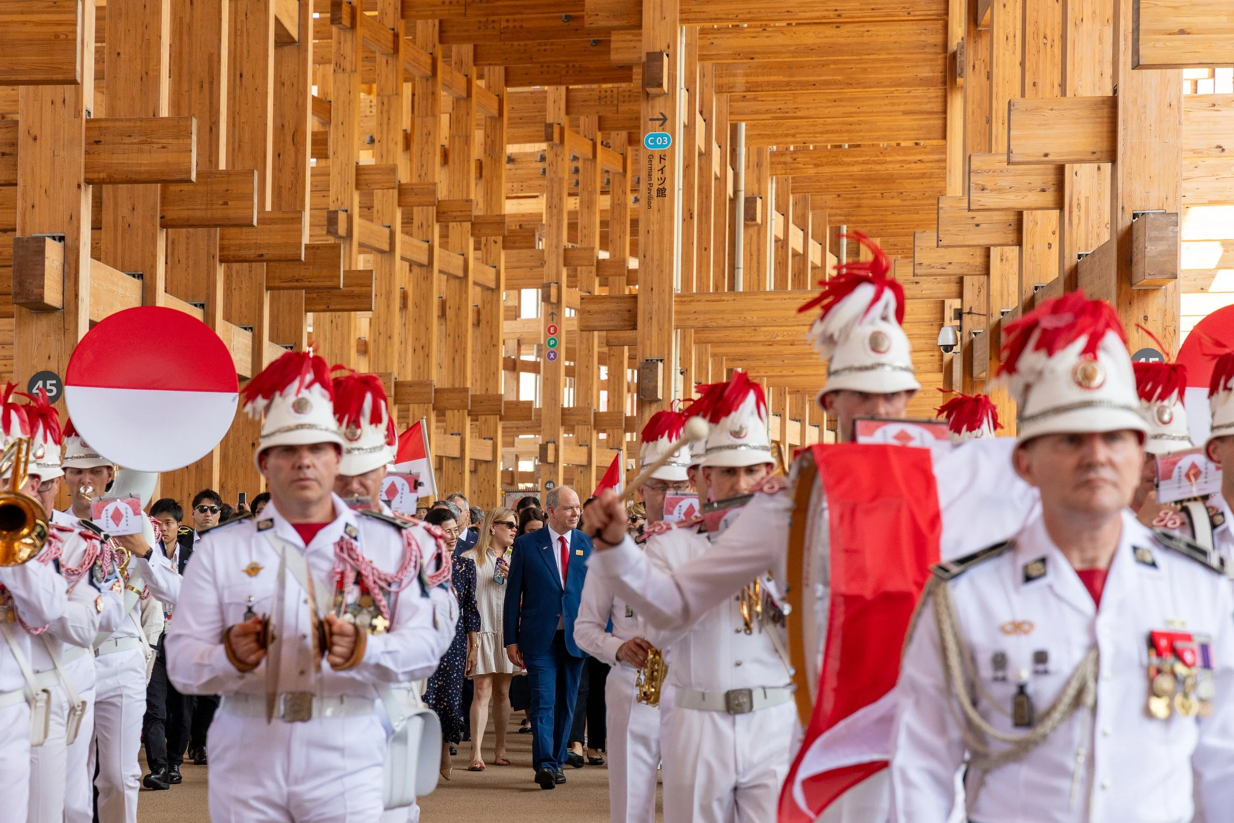 Une procession avec des membres en uniforme blanc avec des chapeaux décorés de plumes rouges, dans un espace en bois avec des drapeaux rouges et blancs, probablement lors d'une cérémonie ou célébration officielle.
