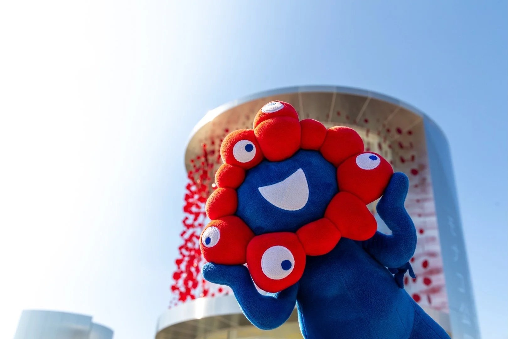 Une main en gant bleu tenant un marionnet de forme ronde avec un visage souriant et entouré de petites créatures rouges avec des yeux blancs, devant un bâtiment moderne avec des arches et des sculptures en forme de petits cercles rouges, sous un ciel bleu clair.