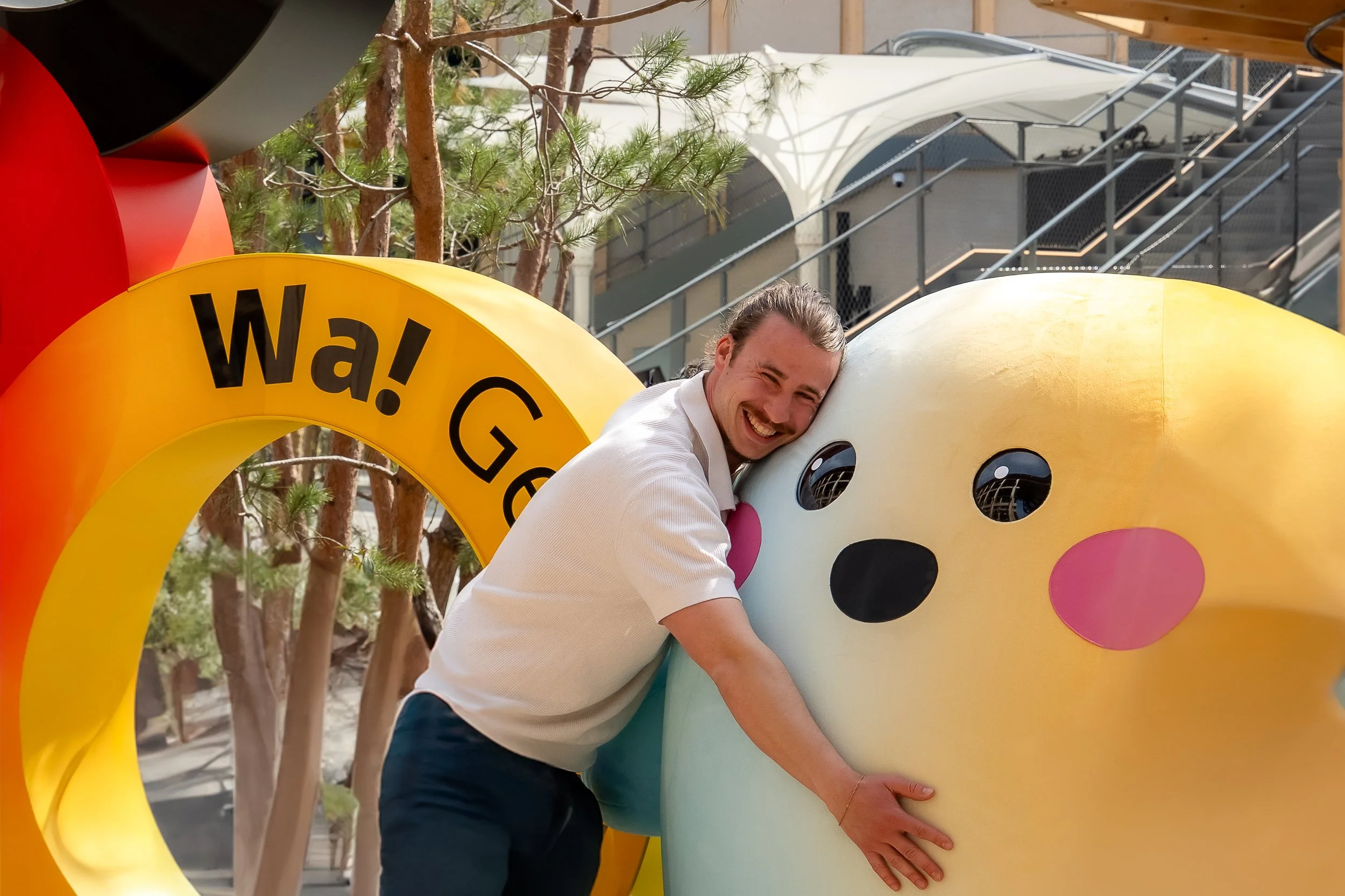 Un homme souriant en polo blanc serre un grand jouet de bébé en forme d'ours blanc avec des yeux noirs et des joues roses, à côté d'une structure colorée avec un arc jaune portant l'inscription "Wa! Ge" dans un espace extérieur avec des arbres et un bâtiment en arrière-plan.