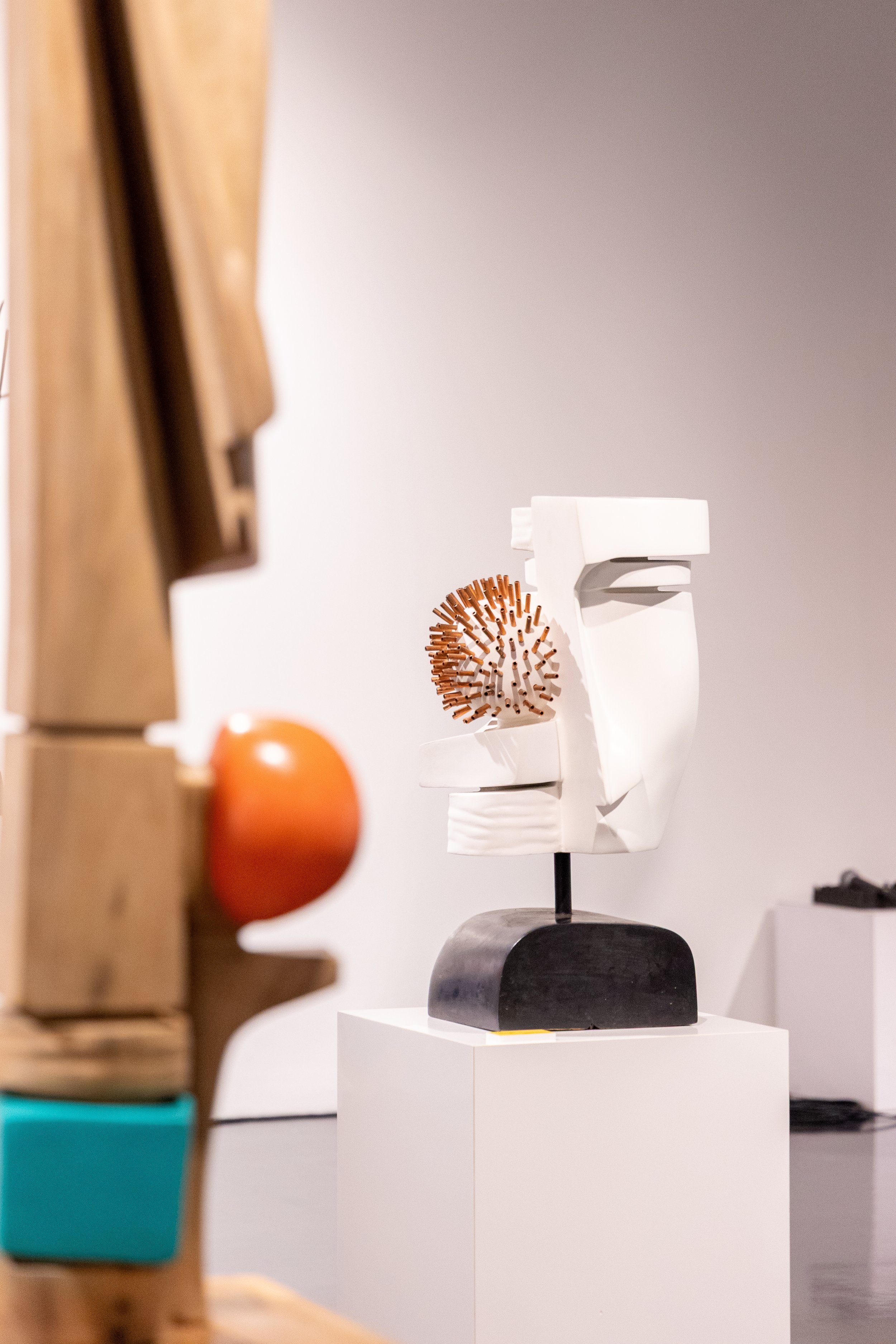 Sculpture moderne en forme de visage abstrait, blanc avec éléments en bois, placée sur un socle noir, exposée dans une galerie d'art.