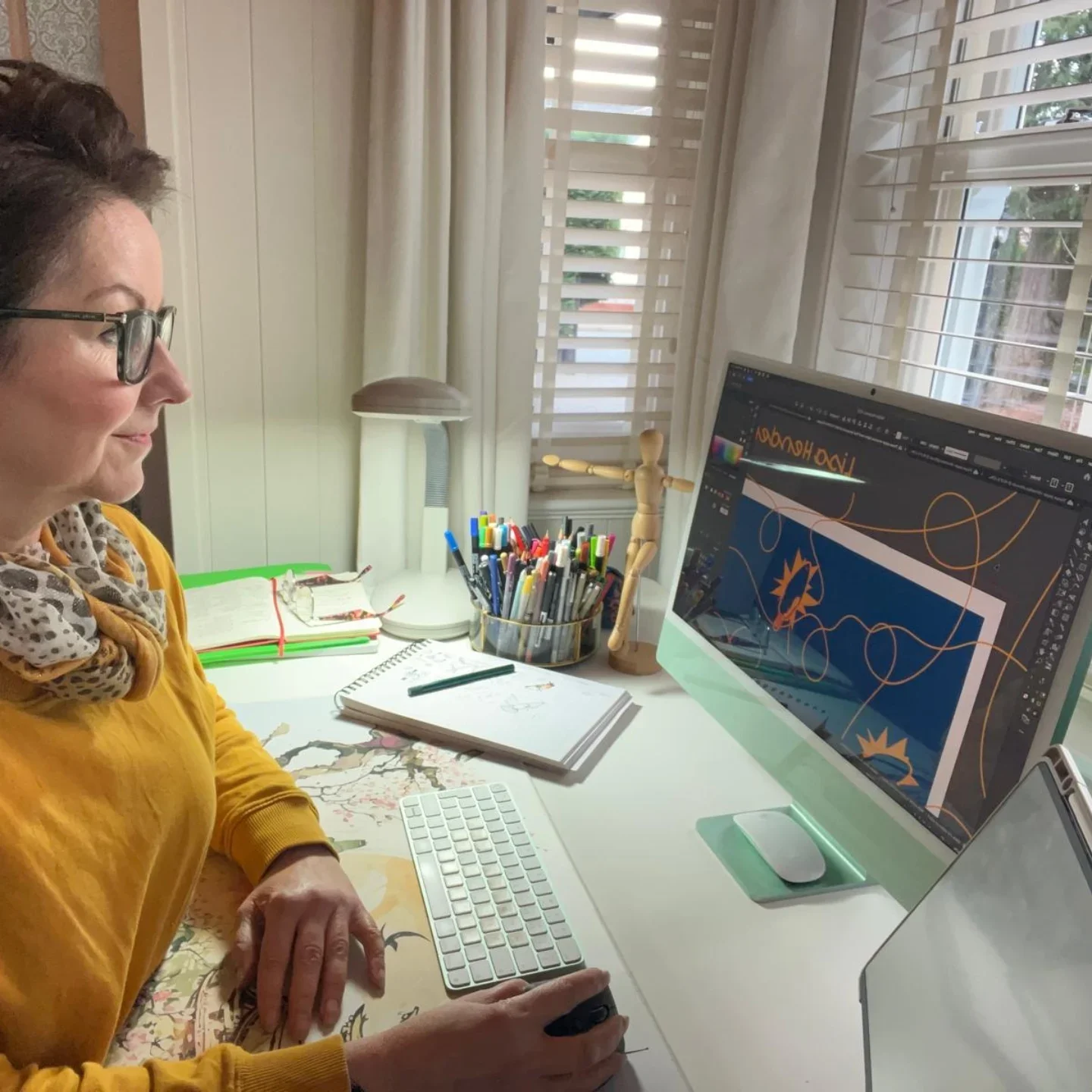 Woman working at a desk with a MAC computer, surrounded by pattern work, notebooks, pens, and a wooden mannequin figurine in their studio