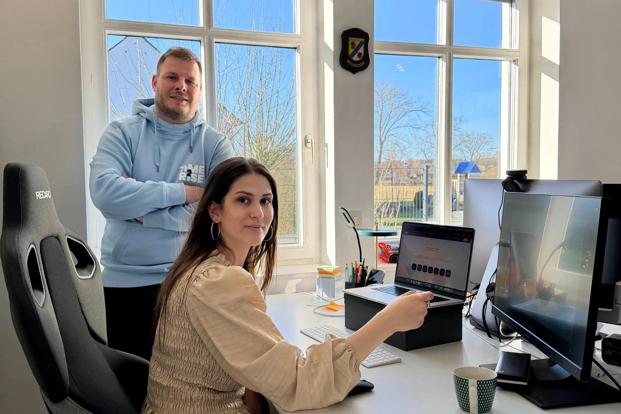 Zwei Personen, eine sitzend an einem Schreibtisch und eine stehend hinter ihr, in einem Büro mit großen Fenstern.