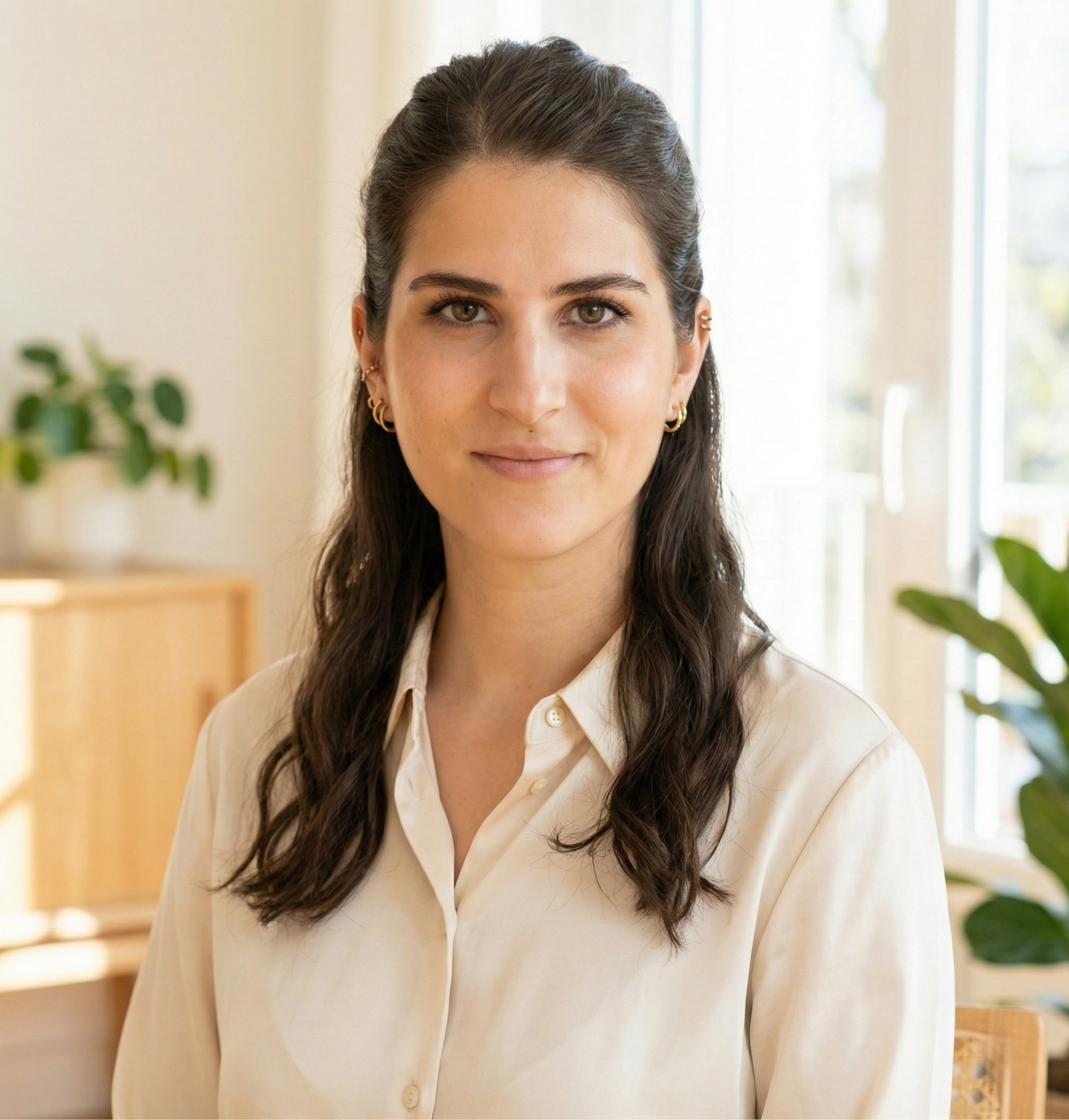 Pia-Teresa Kothe mit dunklen, gewellten Haaren, trägt eine cremefarbene Bluse, sitzt in einem hellen Raum mit Pflanzen im Hintergrund.