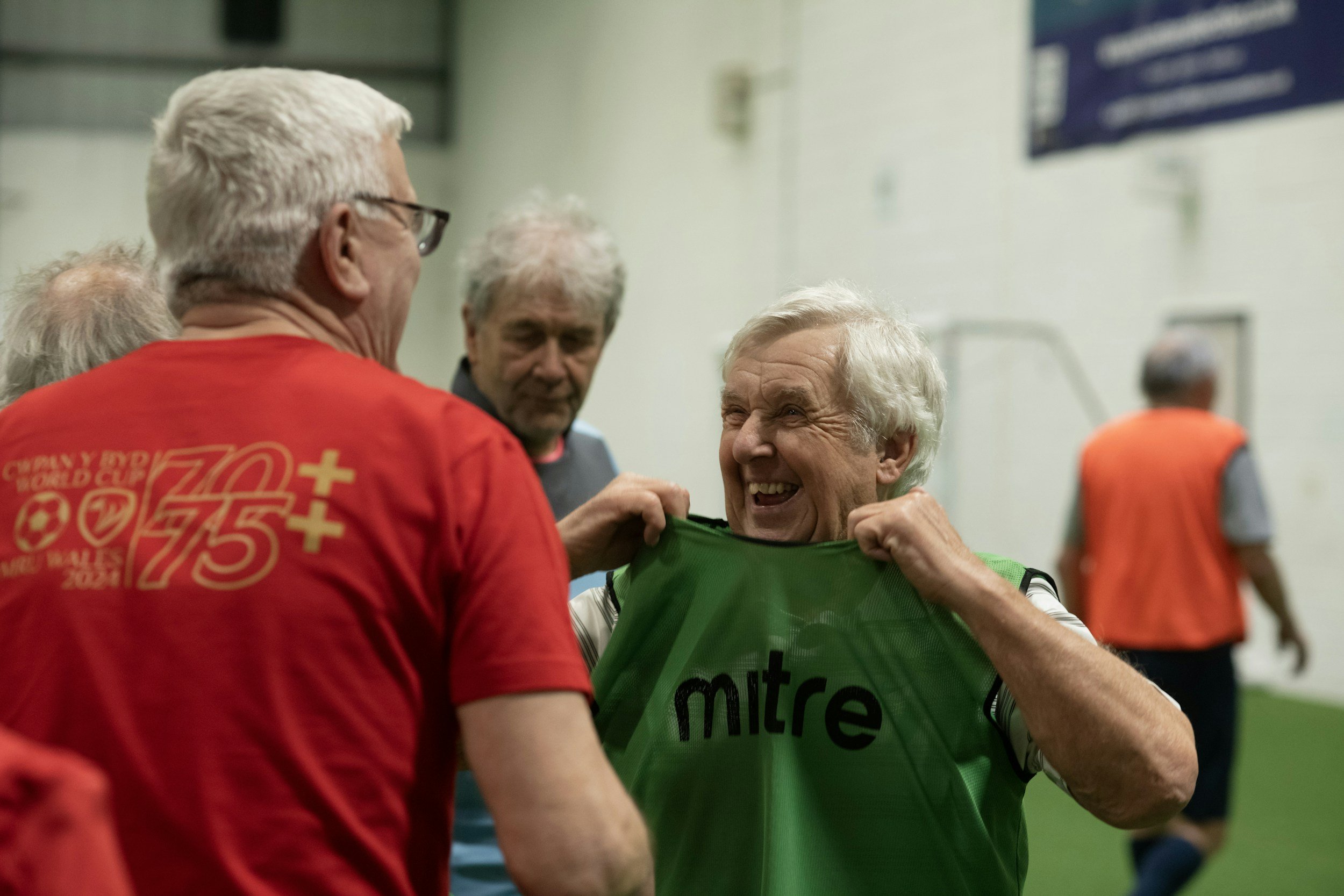 Älterer Mann, lachend und fröhlich, beim Anlegen eines grünen Trikots mit „mitre“ an einem Sportevent, umgeben von weiteren älteren Menschen in einem Indoor-Sportzentrum