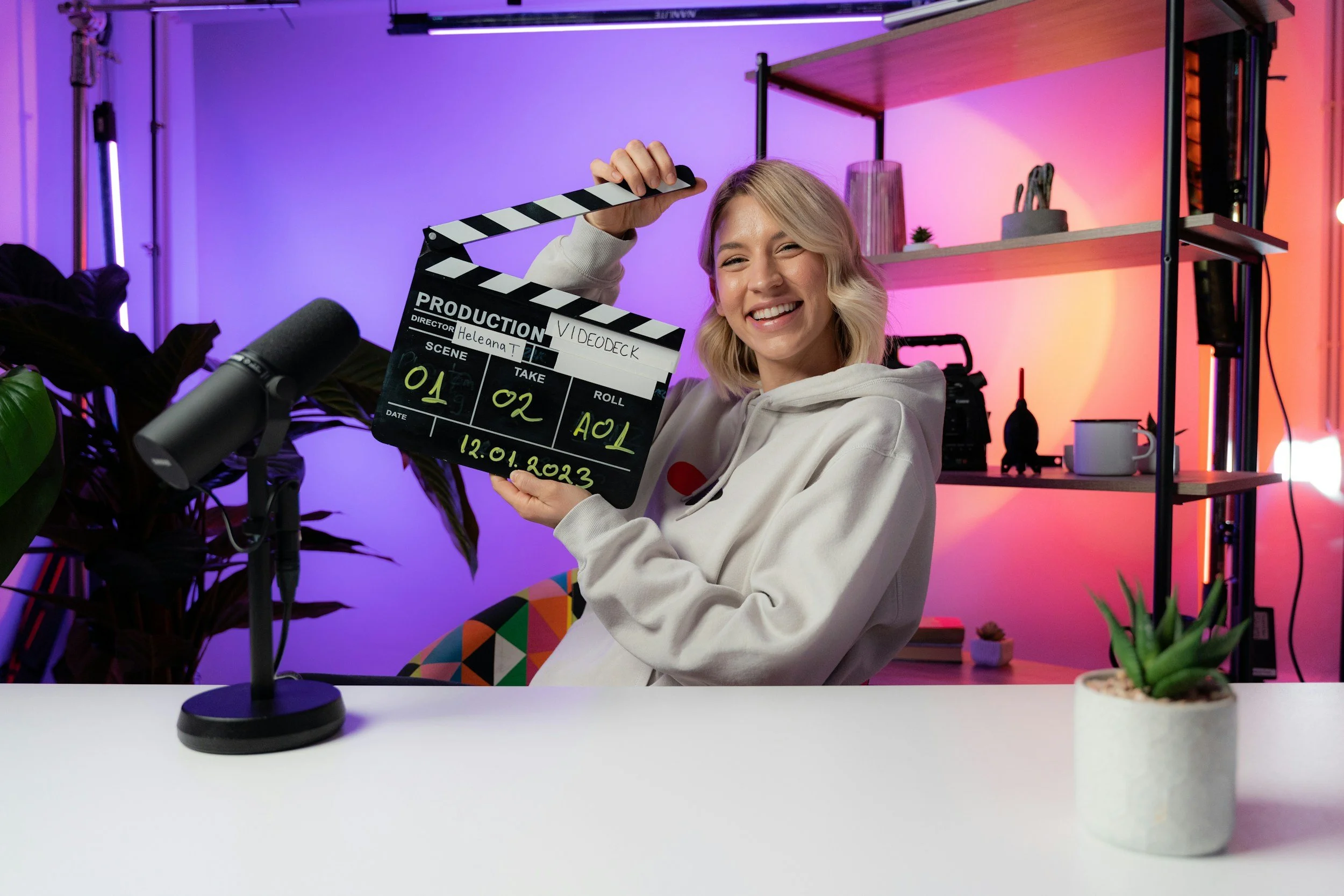 Junge Frau hält eine Filmklappe vor einer bunten, modernen Kulisse mit Pflanzen und Regalen, während sie lächelnd in die Kamera schaut.