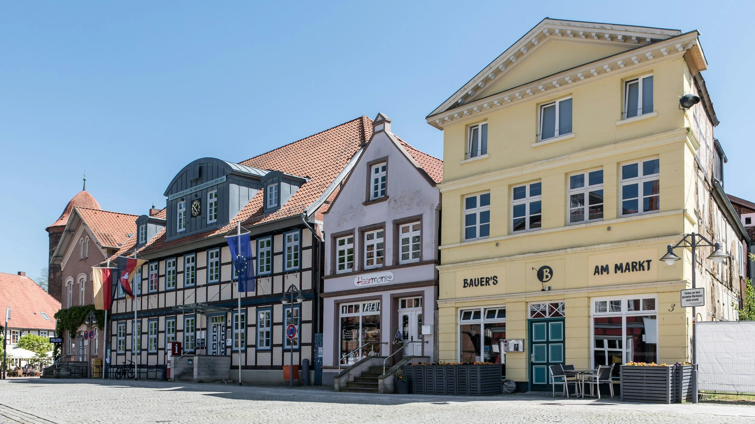 Mehrstöckige historische Gebäude in Lüchow-Dannenberg, darunter ein Fachwerkhaus mit Flaggen, ein gelbes Haus mit Außenbestuhlung, bei strahlendem Himmel.