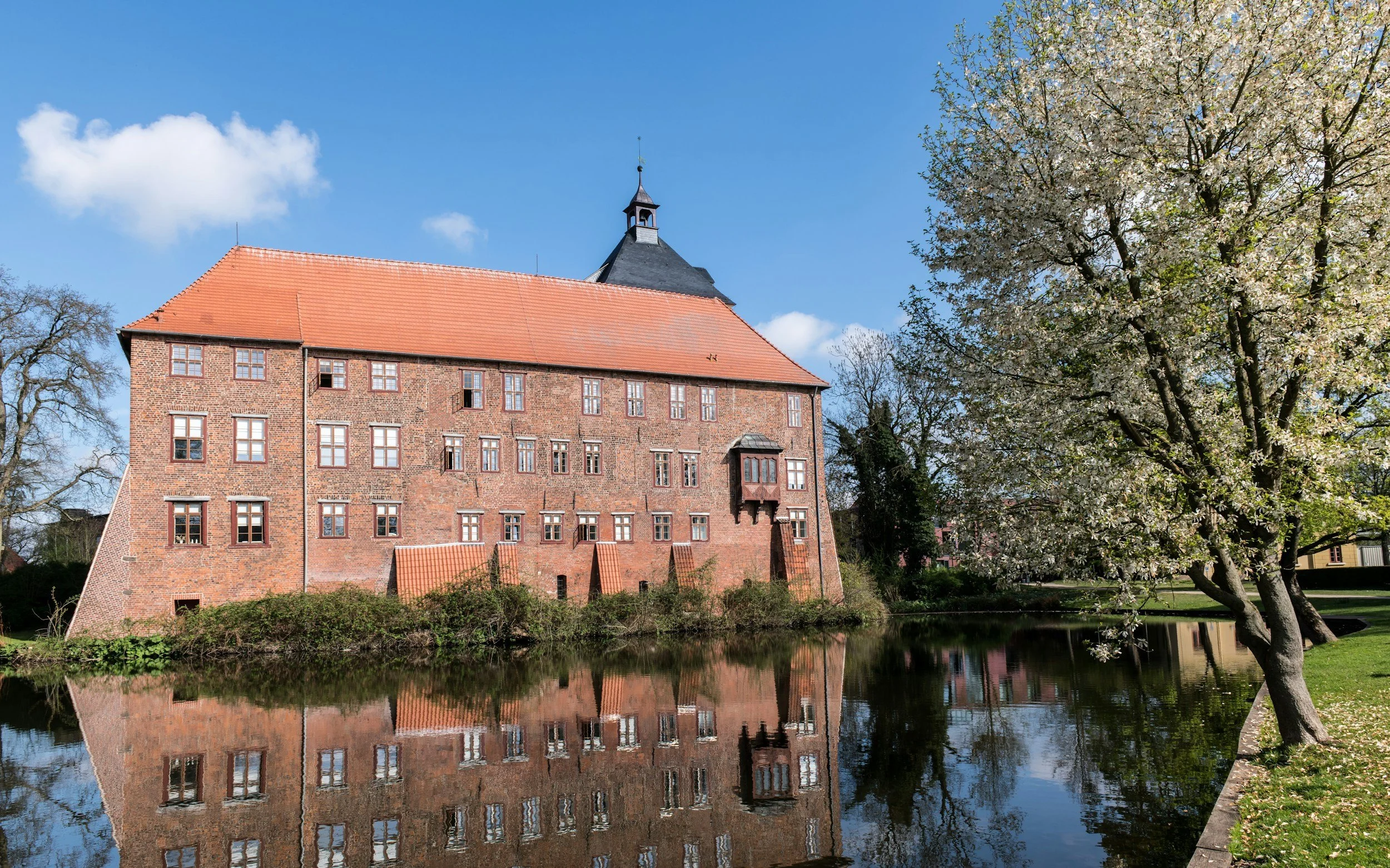 Ein historisches Backsteingebäude mit roten Dachziegeln in Winsen (Luhe), Landkreis Harburg, neben einem Schlossteich, umgeben von Bäumen und einer blühenden Baumkrone im Frühling bei schönem blauen Himmel.