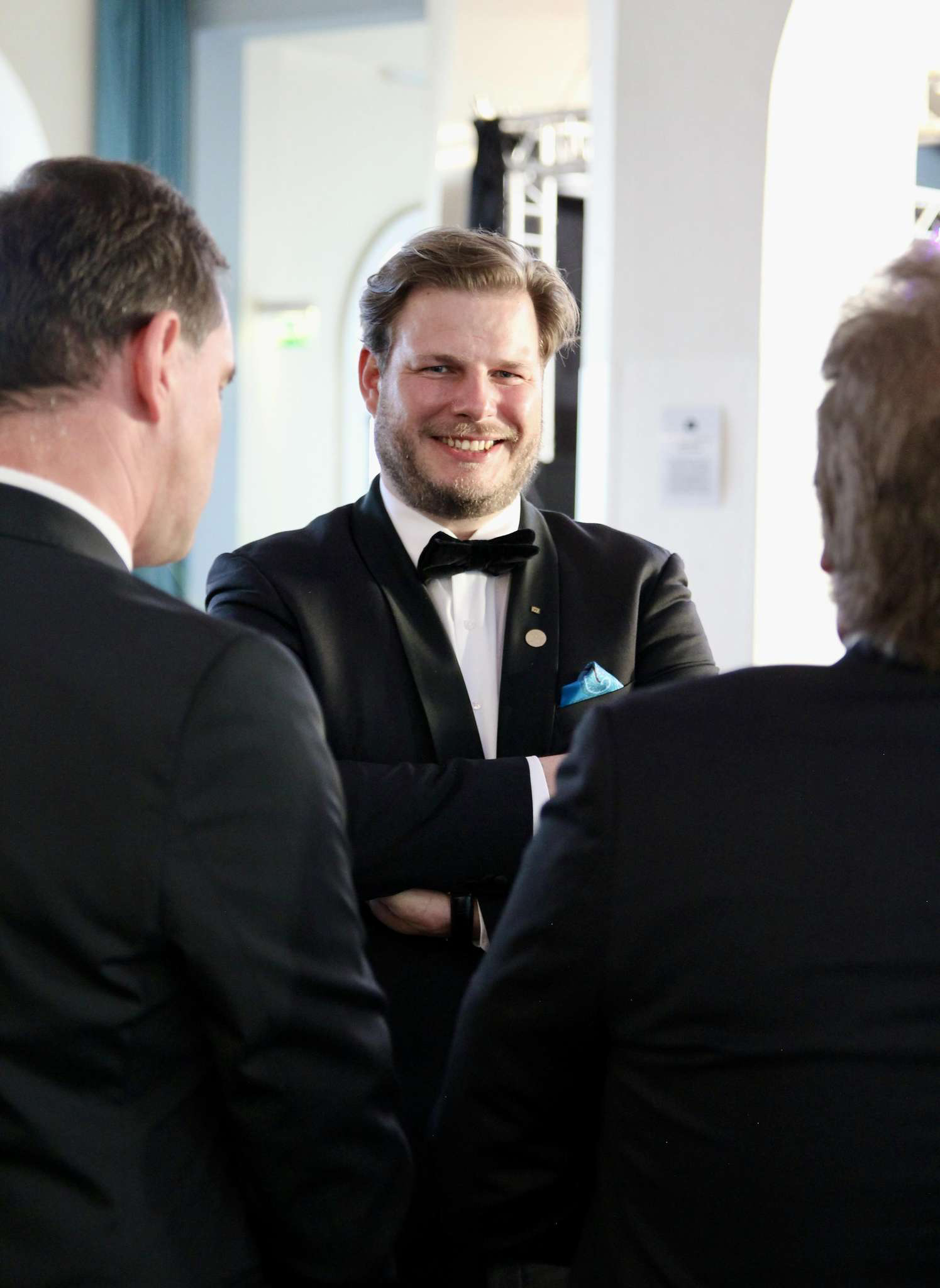 Heiko Kösling. Drei Männer in formeller Kleidung, zwei mit schwarzen Anzügen, unterhalten sich bei einer Veranstaltung, eine Person trägt einen Smoking mit Fliege.