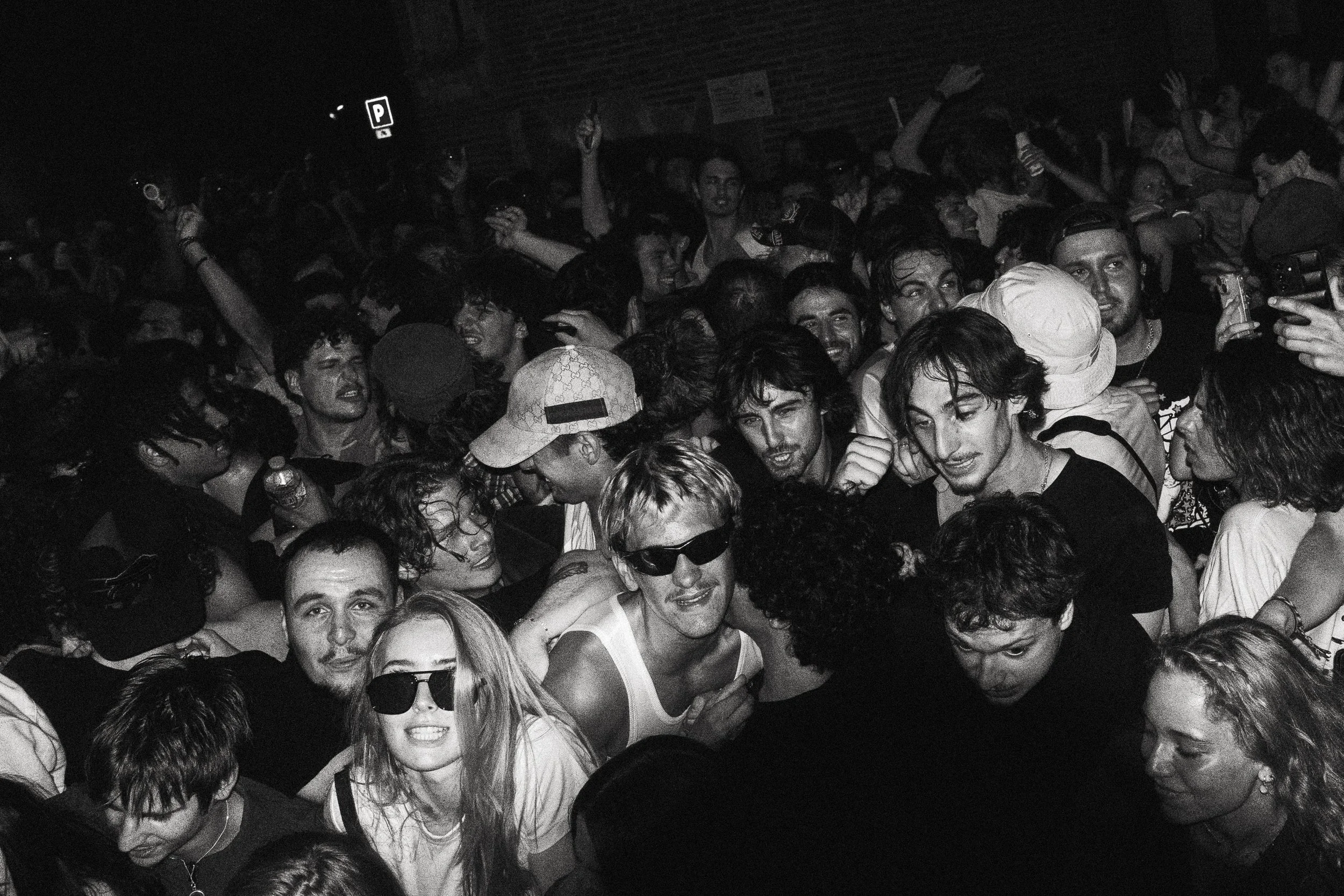 une foule de jeunes gens dans une fête ou un concert, certains portent des lunettes de soleil ou des casquettes, certains prennent des photos ou enregistrent avec leur téléphone portable, ambiance mouvementée et joyeuse