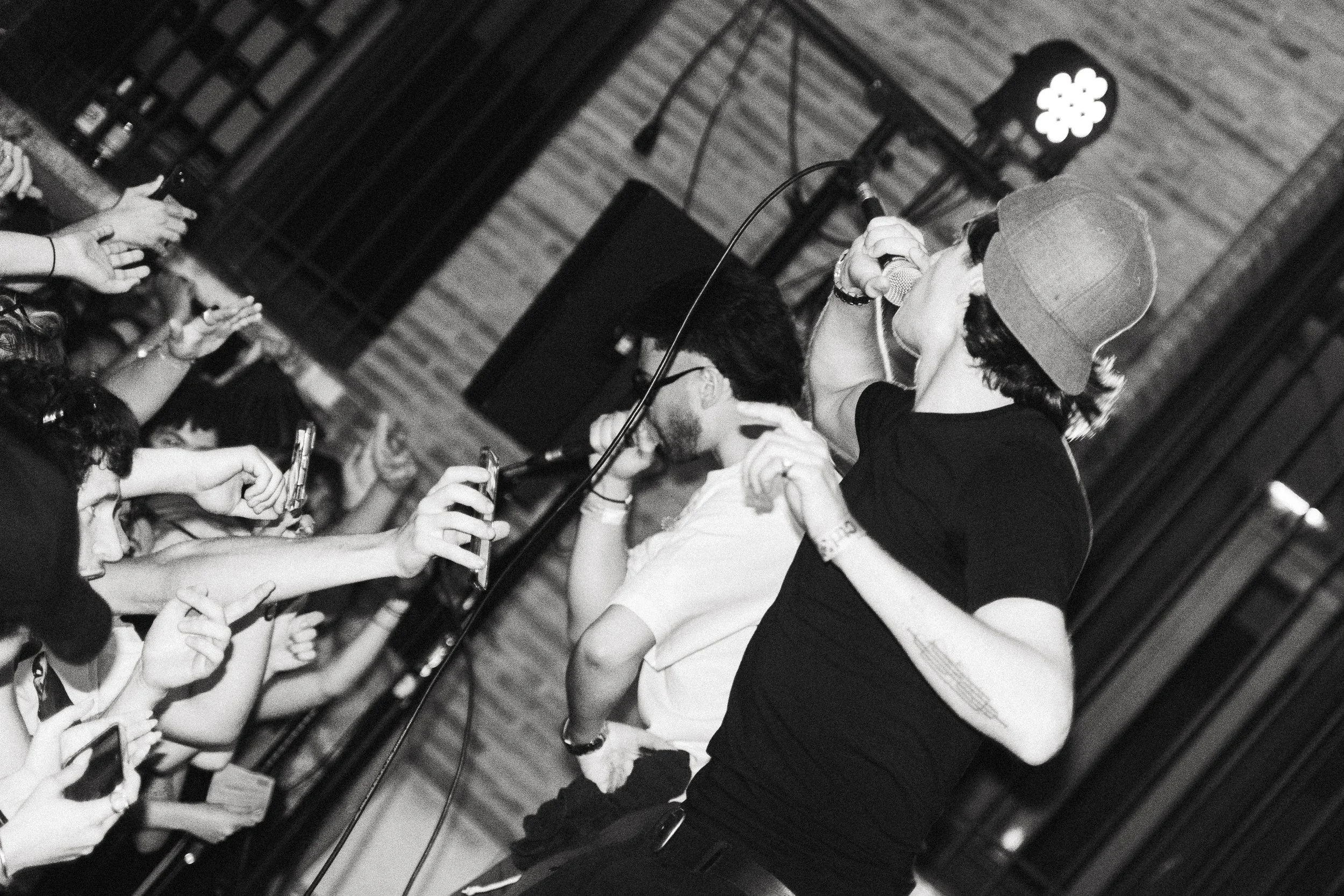Une personne chante dans un microphone lors d'un événement ou concert, entourée de mains de spectateurs qui utilisent leurs téléphones. Fond avec des éléments en bois et un éclairage de scène.