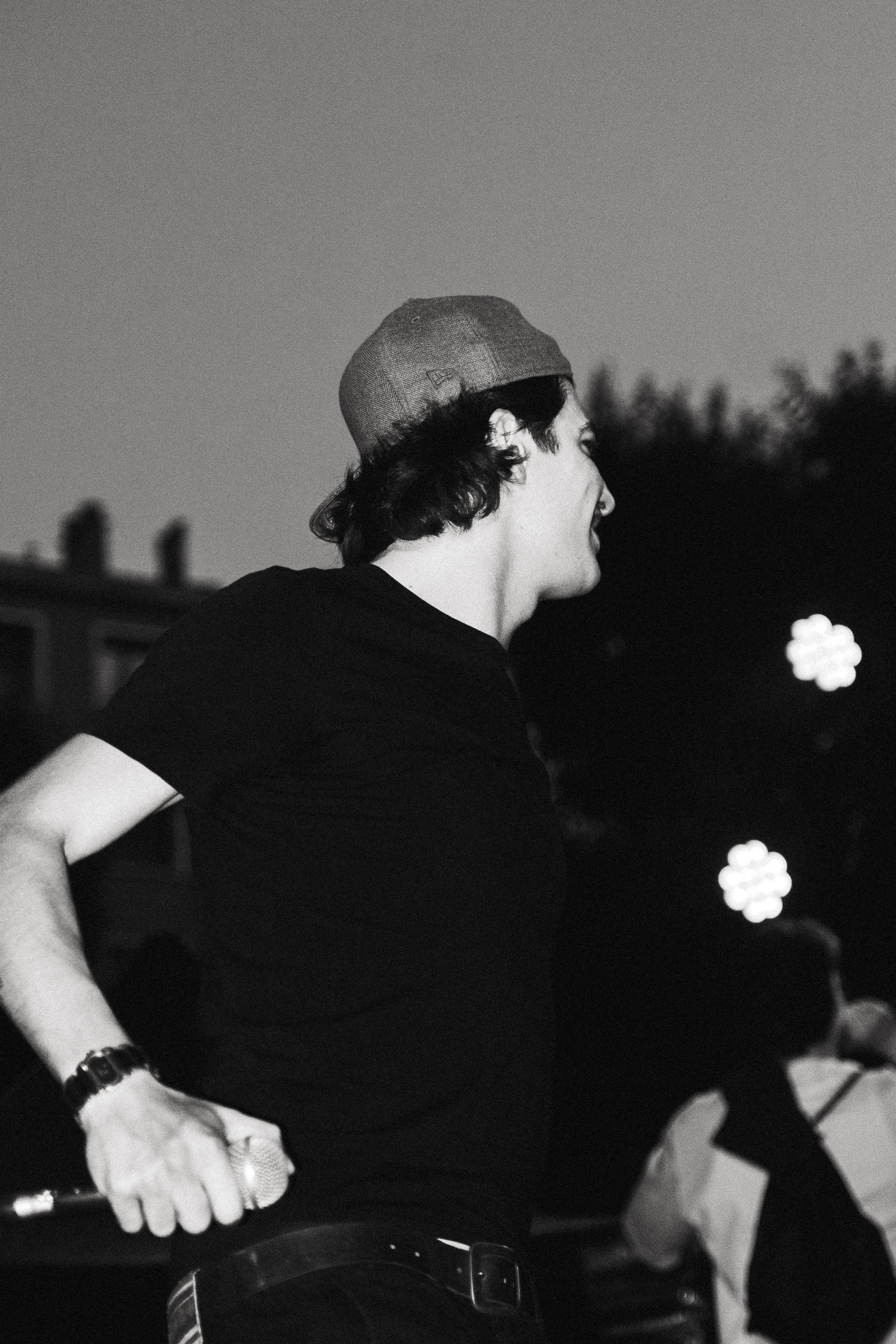 Un homme jeune souriant, portant un t-shirt noir, une casquette grise à l'envers, une montre et une ceinture noire avec une boucle métallique, tient quelque chose dans sa main gauche. Contexte urbain avec des bâtiments et des lumières floues au loin.
