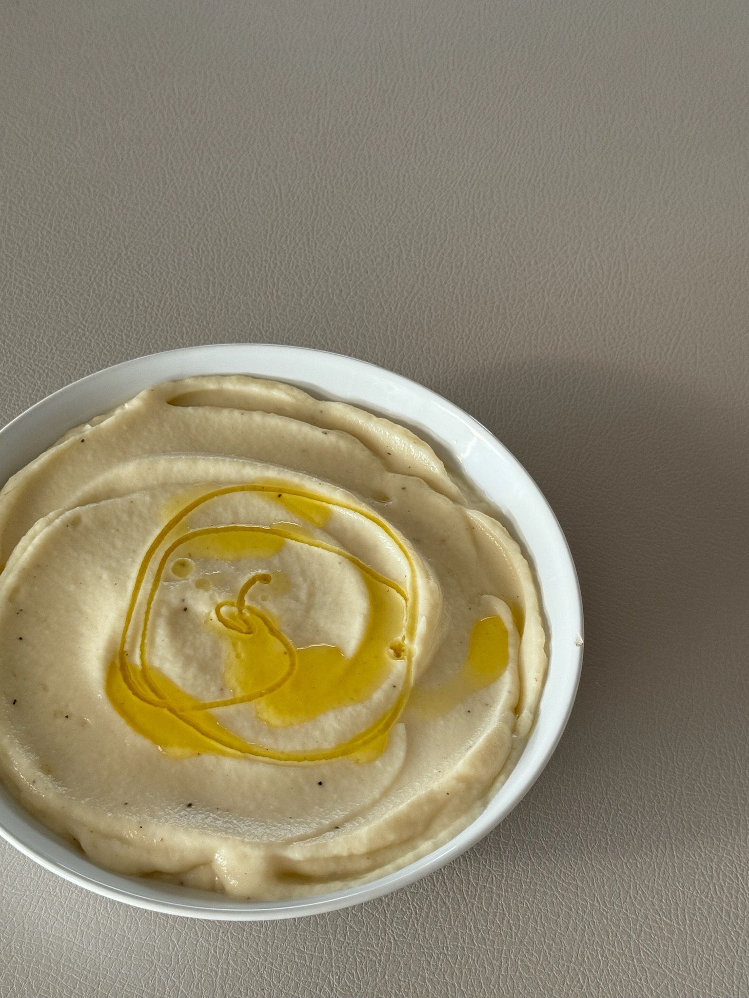 A bowl of creamy mashed potatoes topped with a drizzle of olive oil, placed on a beige surface.